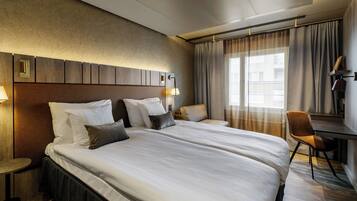 A hotel room with a large bed, a desk, and a chair. There is a window with curtains, a lamp, and a wall-mounted light fixture.