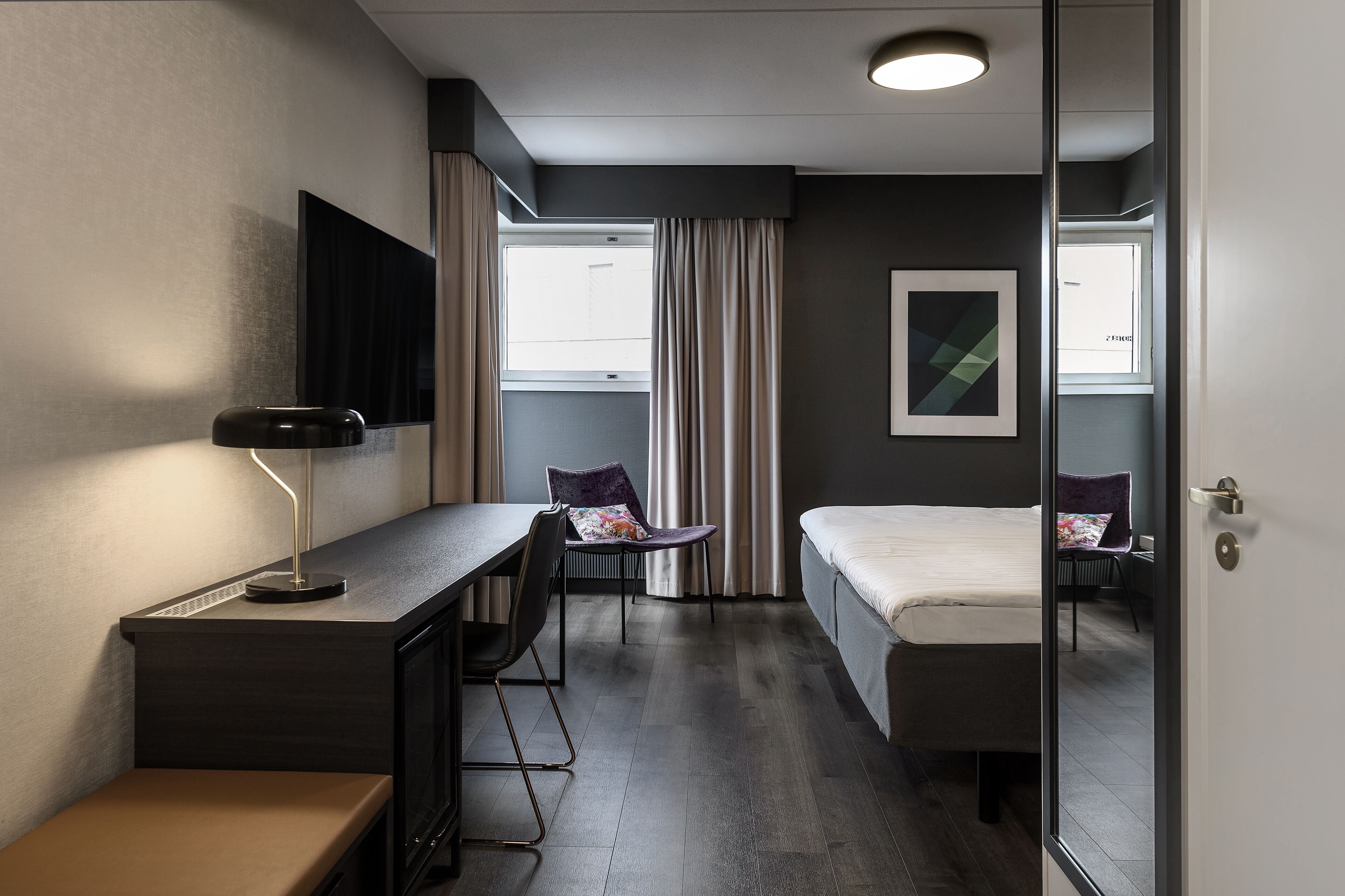 A modern hotel room with a desk, chair, and bed. There is a window with curtains, a flat-screen TV mounted on the wall, and a framed artwork on the wall.