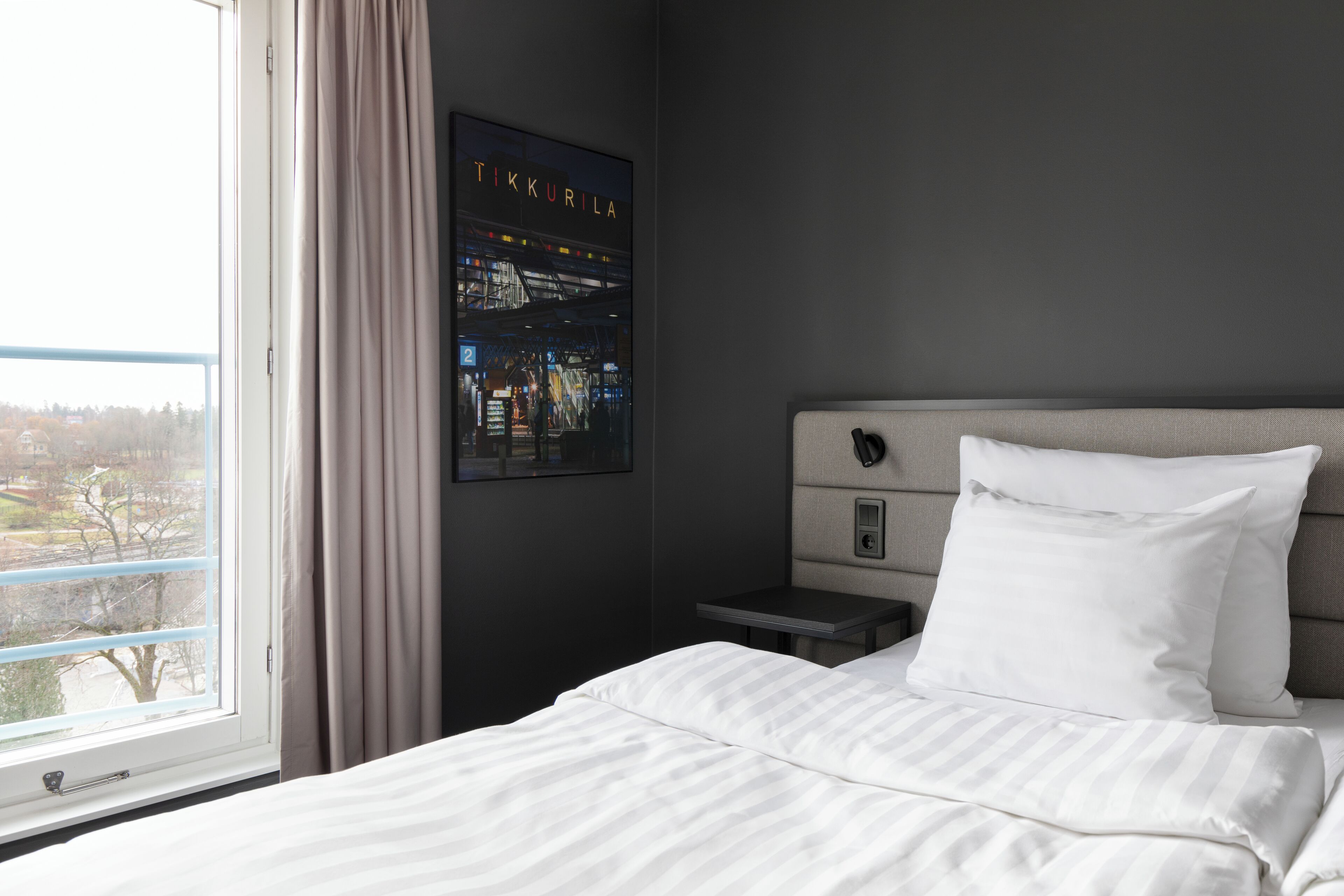 A hotel room with a bed, a nightstand, a wall-mounted poster, and a window with a view of the outside.