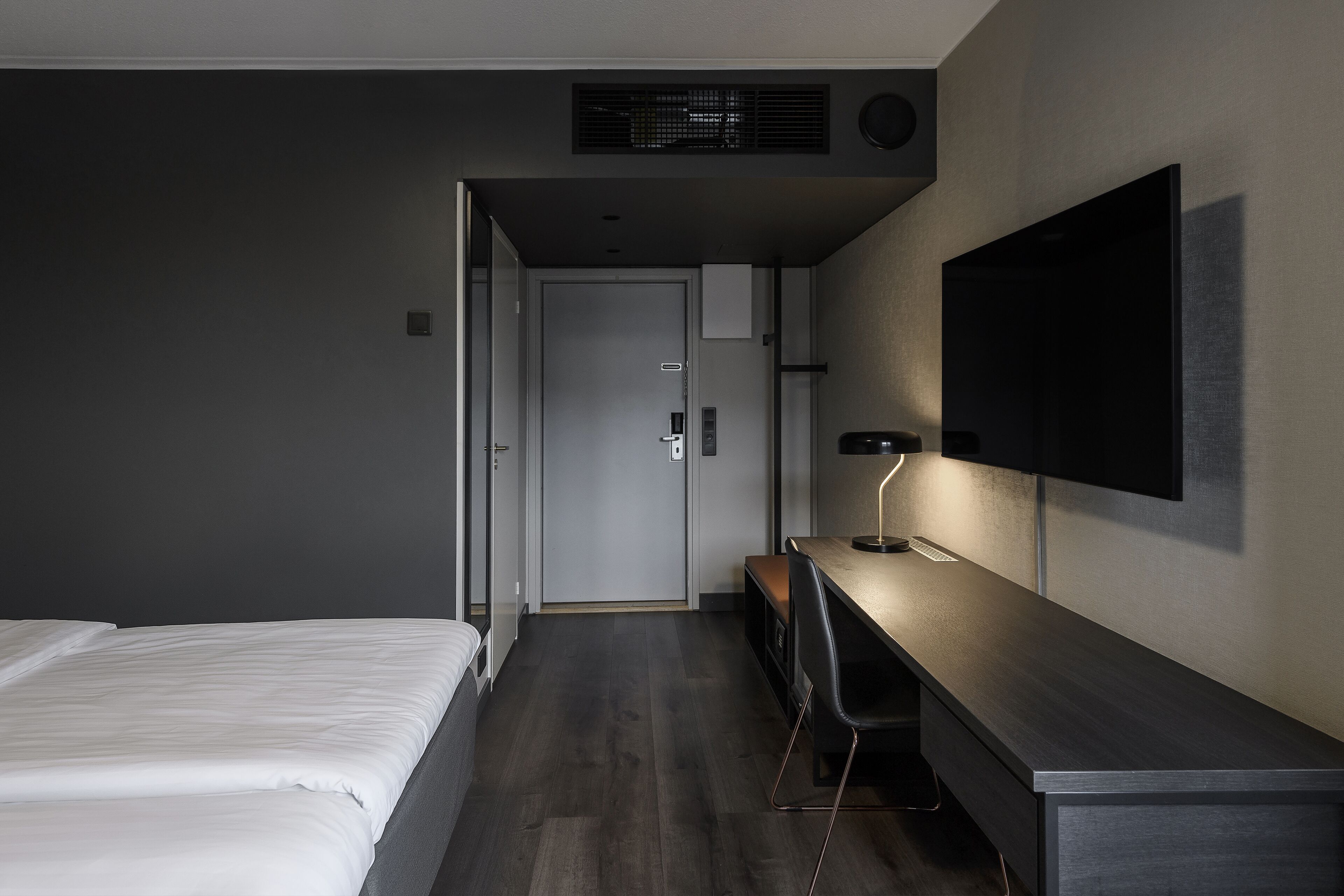 Una habitación de hotel moderna con una cama, un escritorio con una silla, un televisor de pantalla plana montado en la pared y una puerta que da al baño.