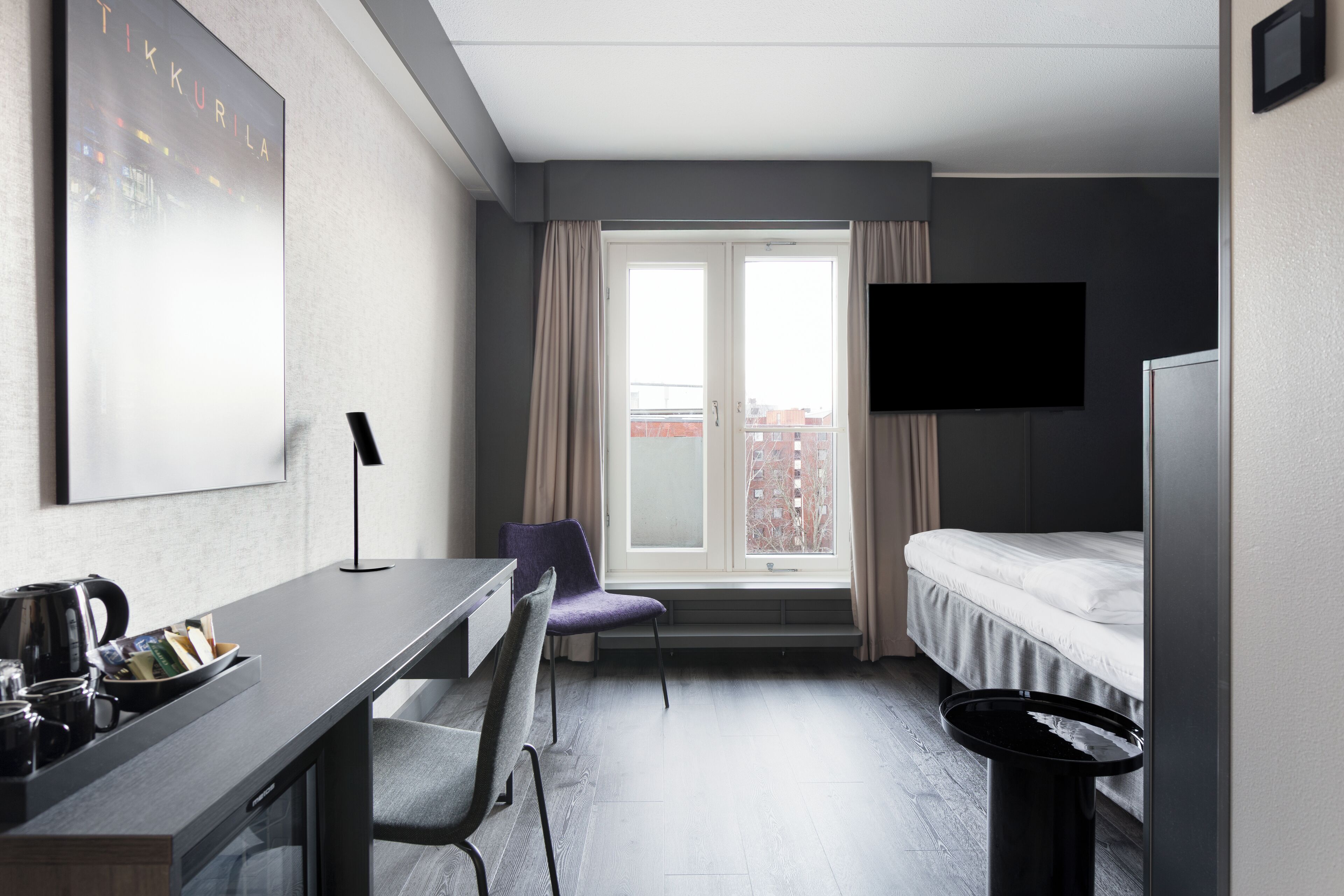 Habitación de hotel con cama, escritorio, silla, televisor, ventana con cortinas y vistas a edificios.