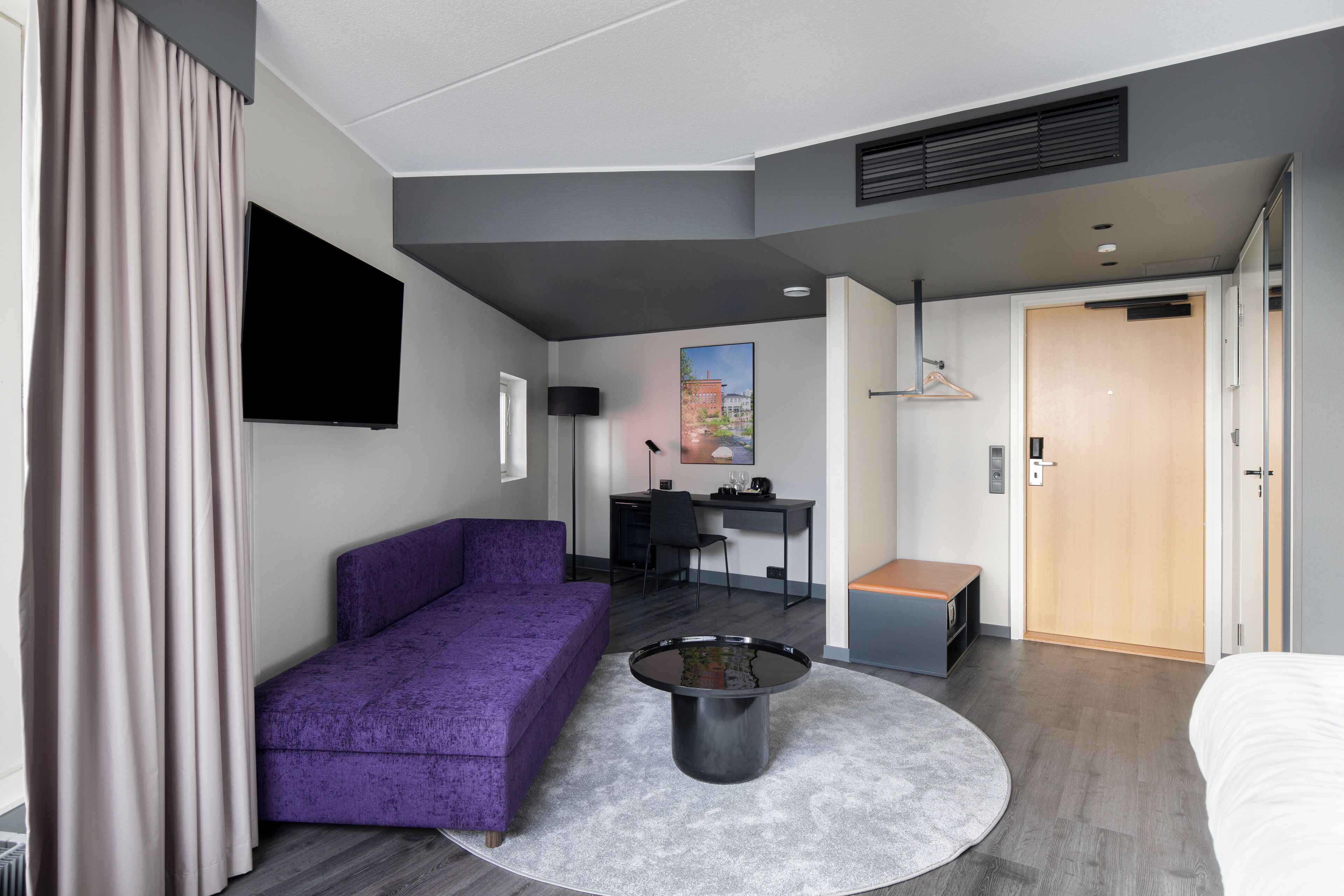 A modern hotel room with a purple sofa, a round coffee table, a desk, and a bed.