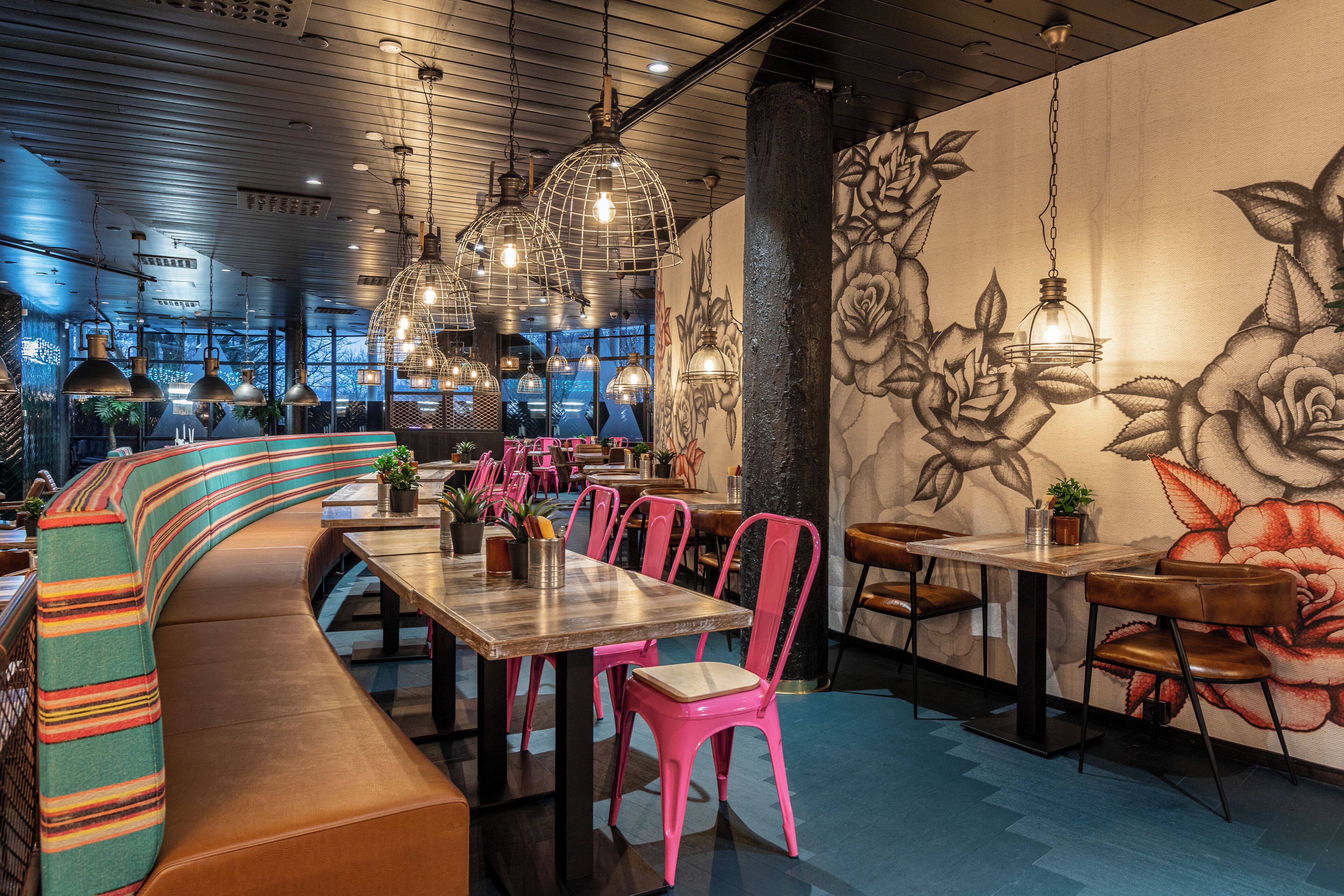 A restaurant interior with a curved bench, tables, pink chairs, and hanging light fixtures.