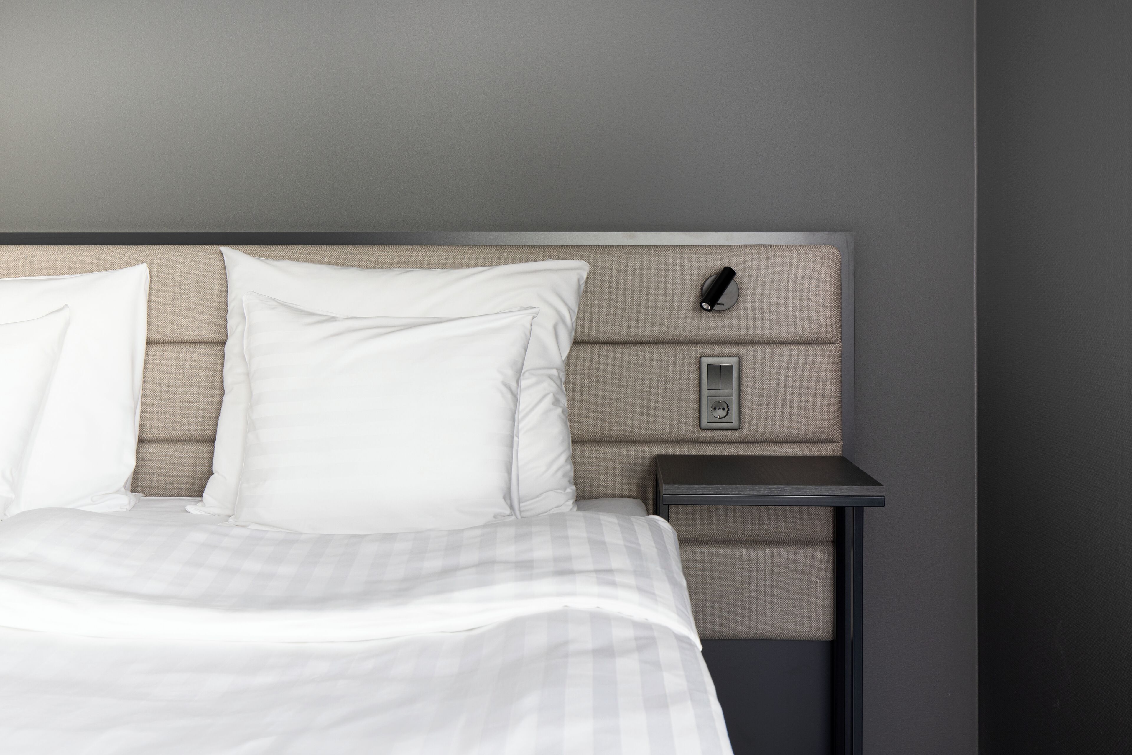 A neatly made bed with white linens, a headboard with a light fabric, and a small bedside table with a power outlet.