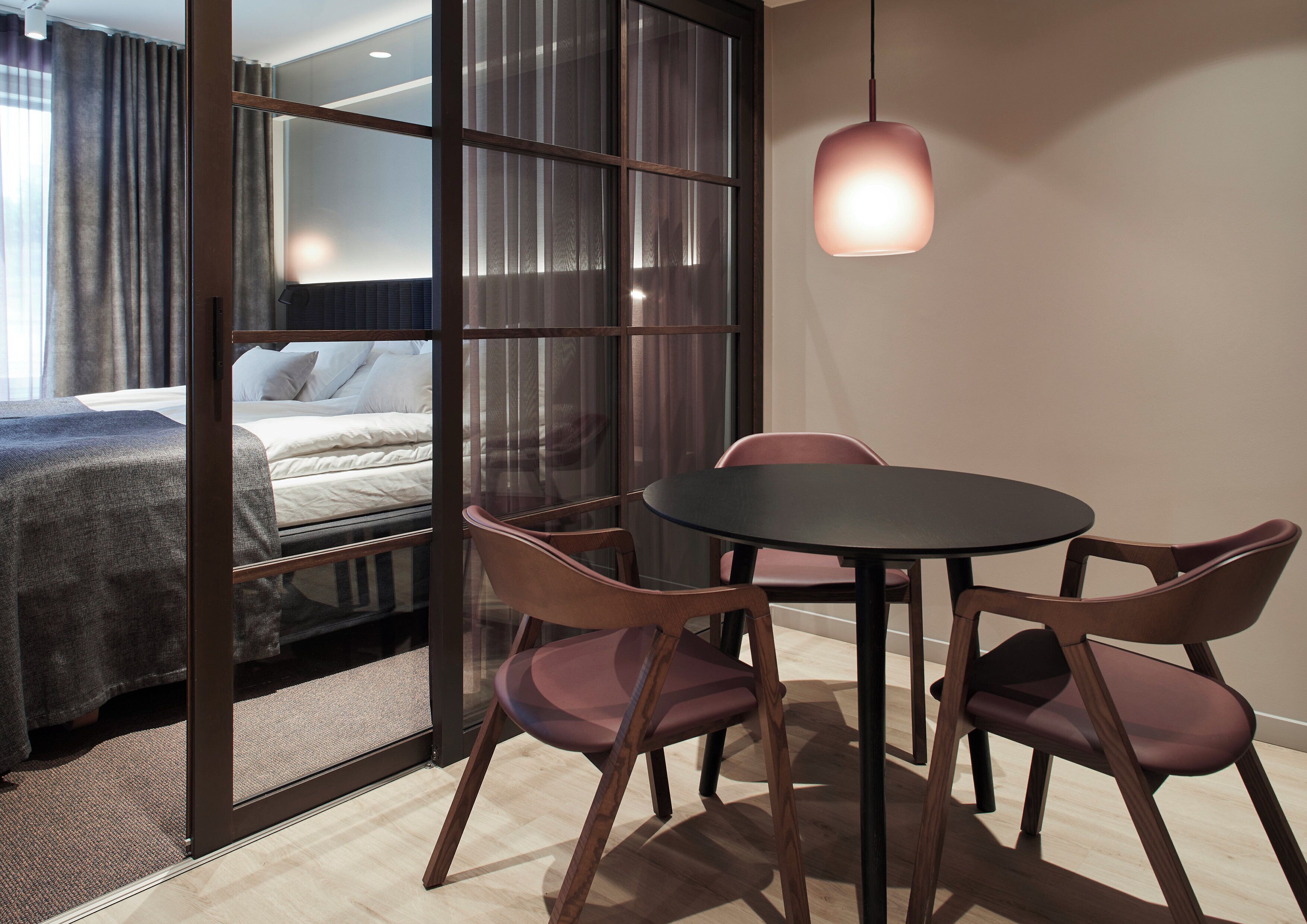 A hotel room with a bed, a dining table with chairs, a pendant light, and a view through a glass partition.