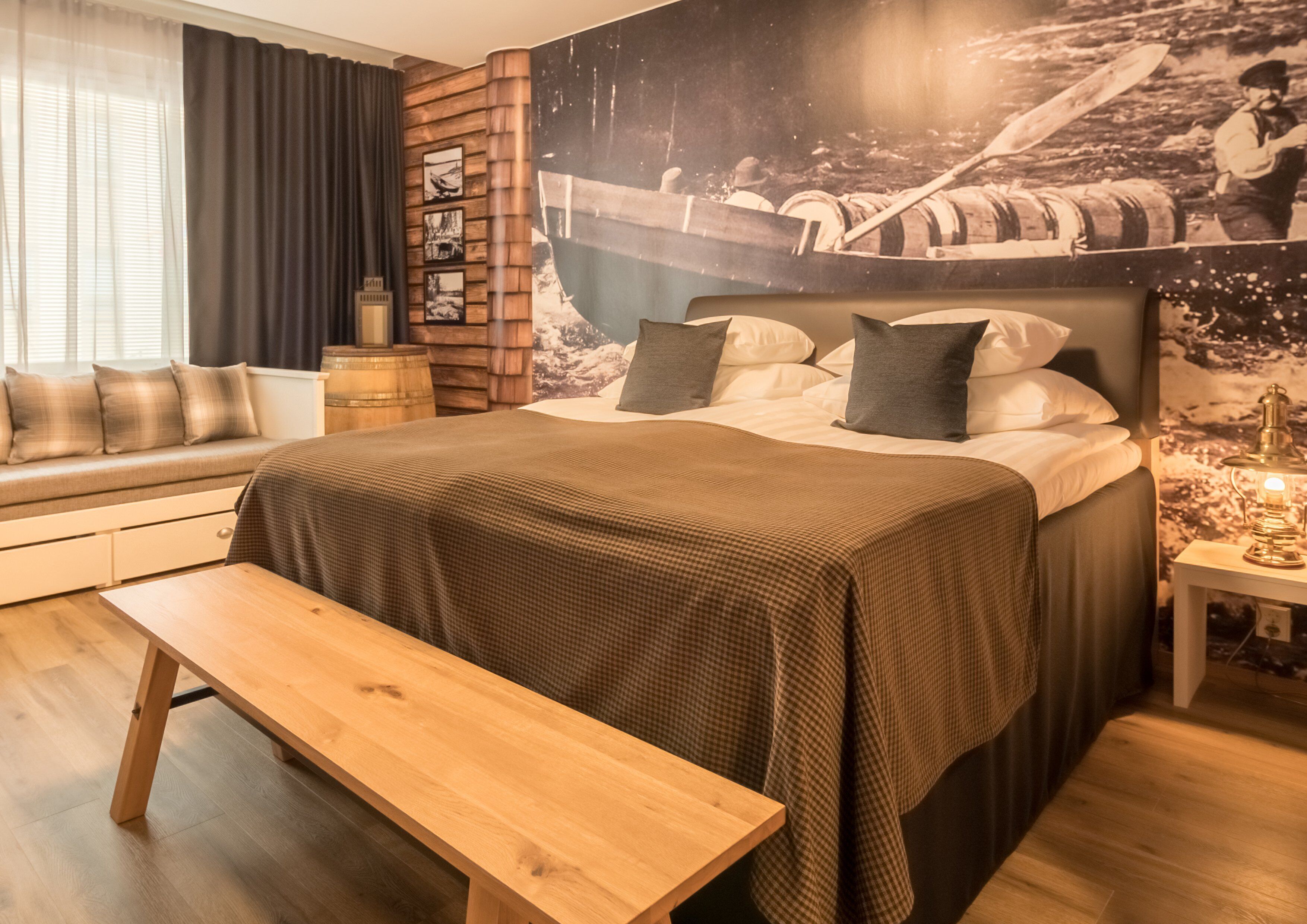 Een slaapkamer met een groot bed, een bankje, een houten hoofdbord, een muurschildering, een raam met gordijnen en een nachtkastje met een lamp.