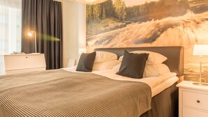 Una habitación de hotel con una cama grande, un cabecero con un cuadro, una mesita de noche con una lámpara y una ventana con cortinas.