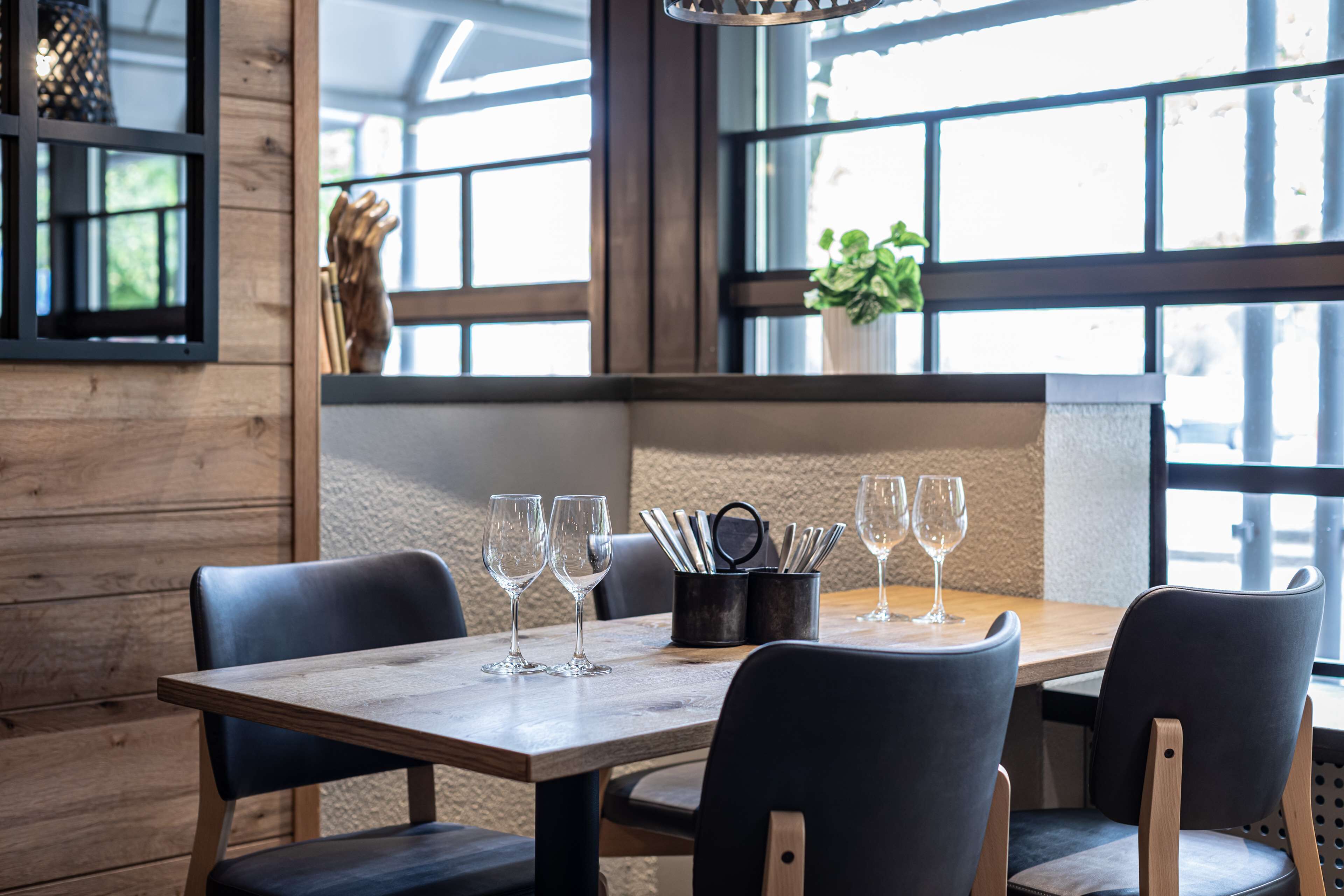 Een restaurantinterieur met een houten tafel, zwarte stoelen en een raam met uitzicht op groen.