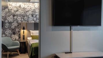 A modern hotel room with a flat-screen TV mounted on the wall, a small bedside table with a lamp, a green chair, and a bed with a green blanket.