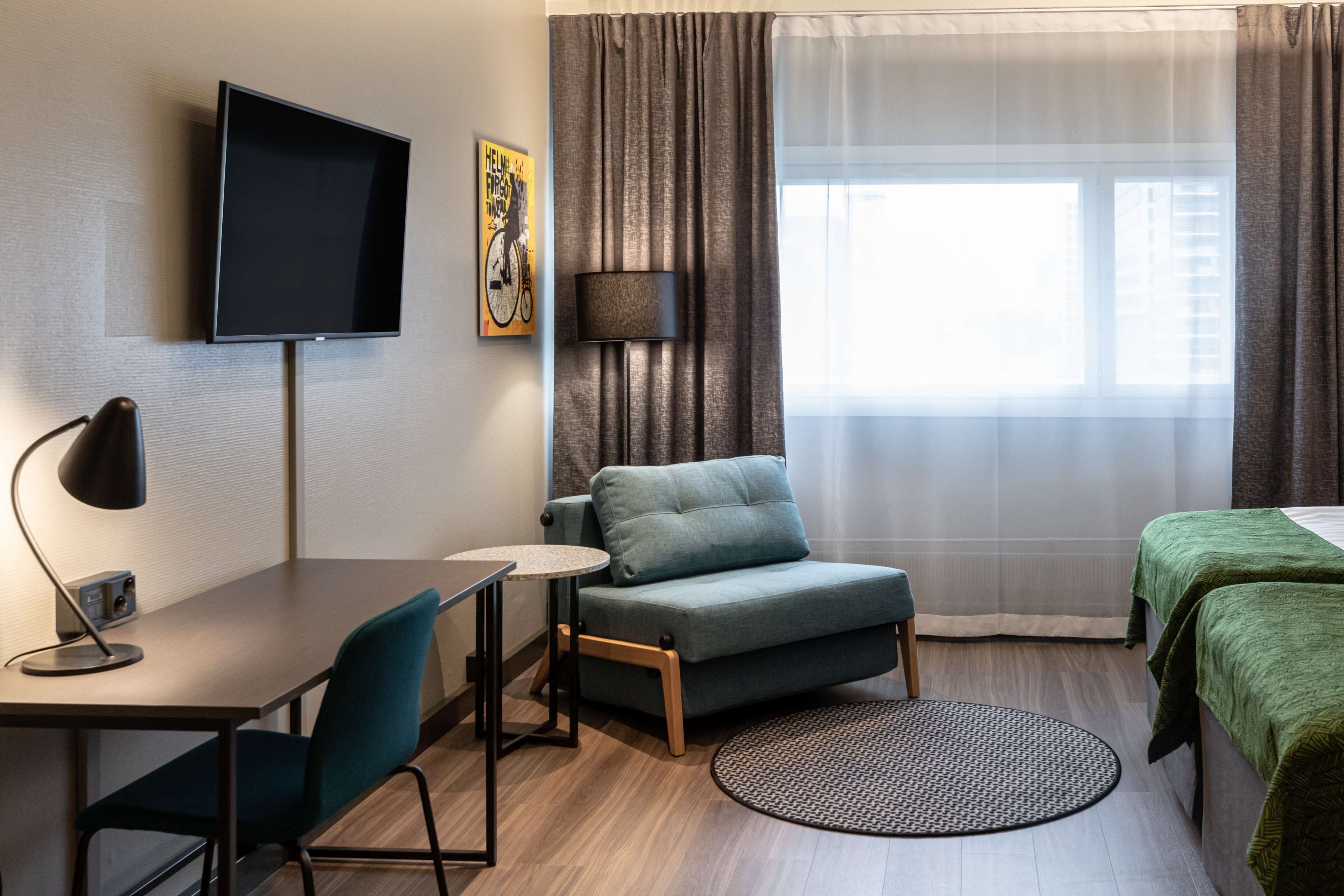 Ein Hotelzimmer mit Flachbildfernseher, Schreibtisch mit Lampe, Sofa, Bett mit grüner Bettwäsche und Fenster mit durchsichtigen Vorhängen.