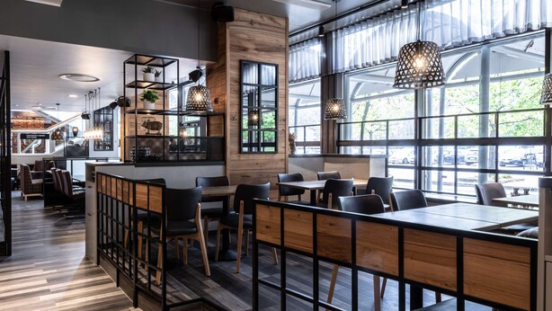 A modern cafe with wooden tables and chairs, large windows, and hanging pendant lights. - Scandic Pasila (Helsinki)