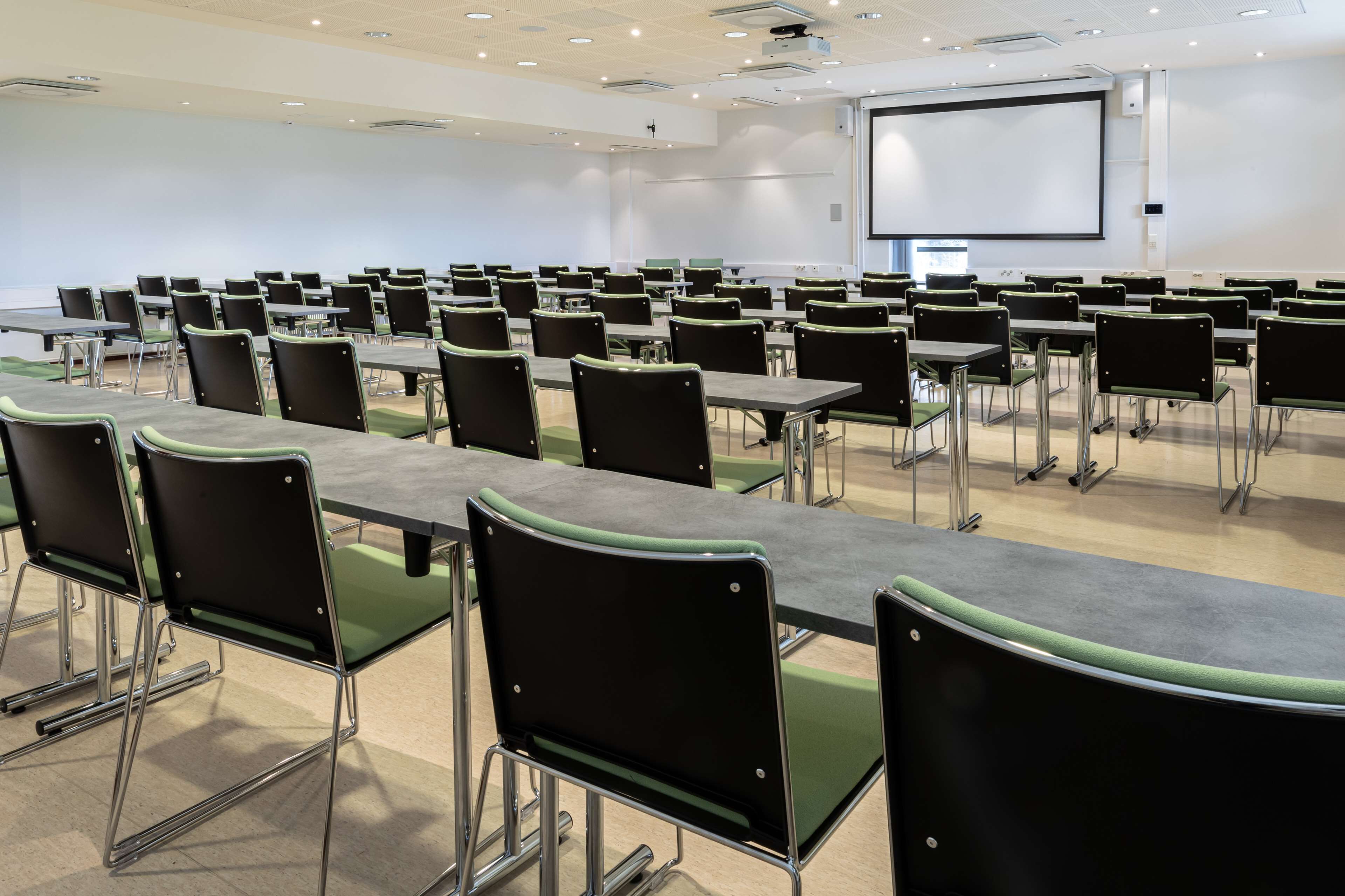 Una sala de conferències amb files de taules i cadires, una pantalla de projector i una pissarra.