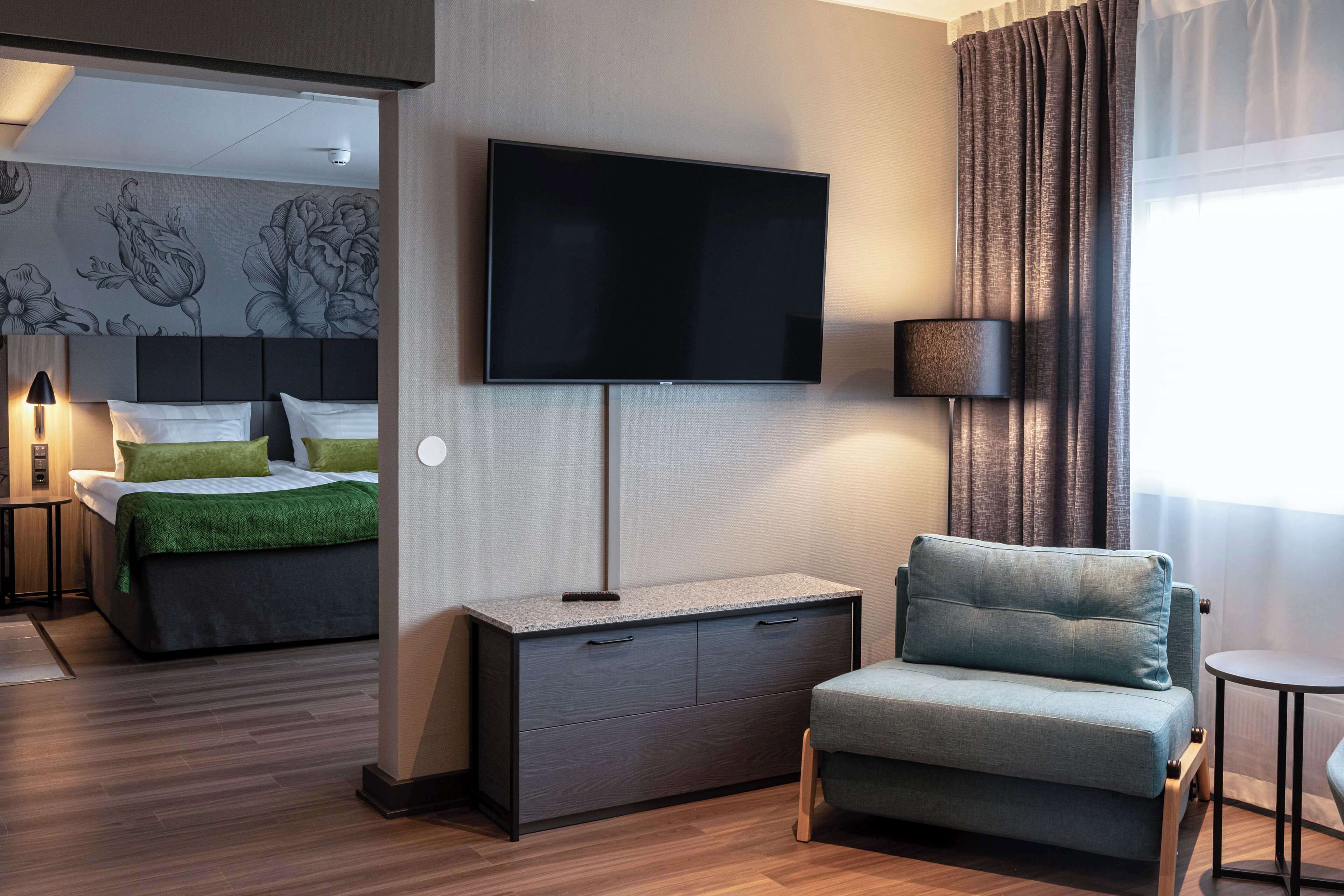 Ett modernt hotellrum med en platt-TV som är monterad på väggen, en säng med gröna kuddar, en grå soffa och ett litet bord med en lampa.