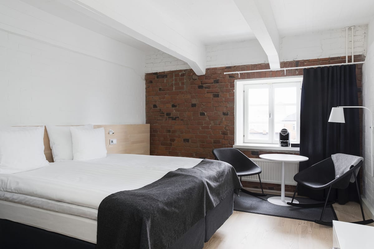 Habitación de hotel con cama, una mesita, dos sillas, ventana con cortinas y pared de ladrillo.