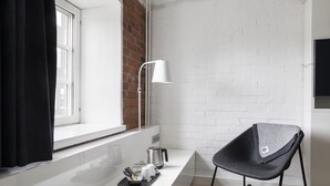 Una habitación moderna con pared de ladrillo blanco, ventana blanca con cortinas negras, escritorio blanco con hervidor, una silla y una lámpara de pie blanca.