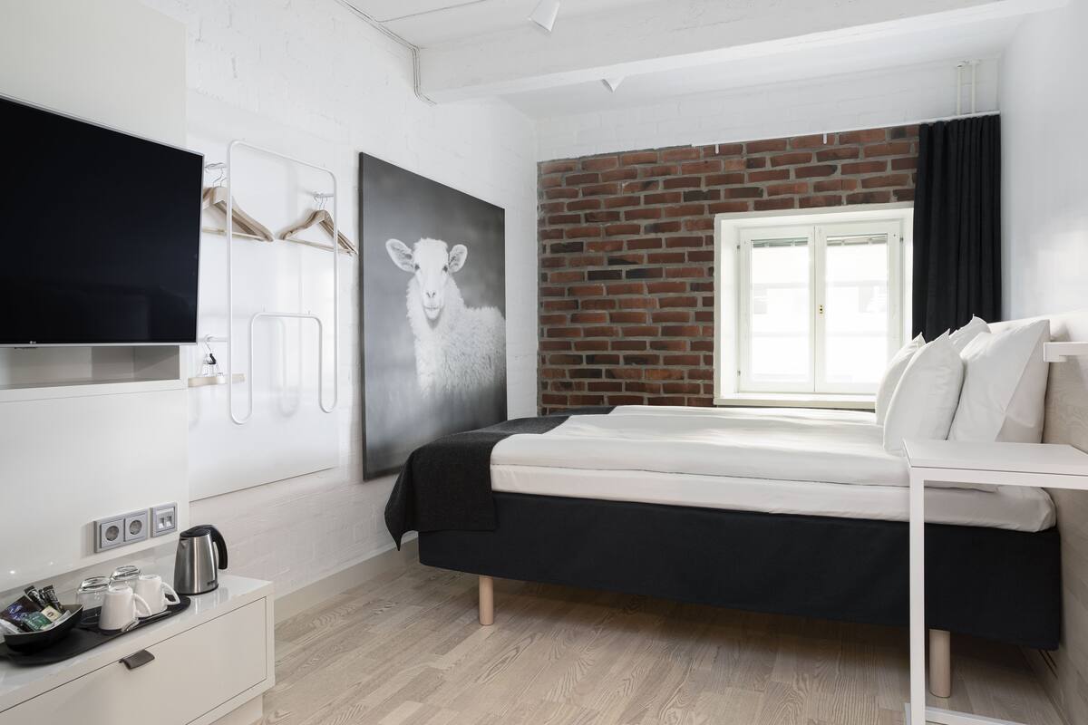 Habitación de hotel moderna con cama, televisión de pantalla plana, cafetera y pared de ladrillo decorativa.