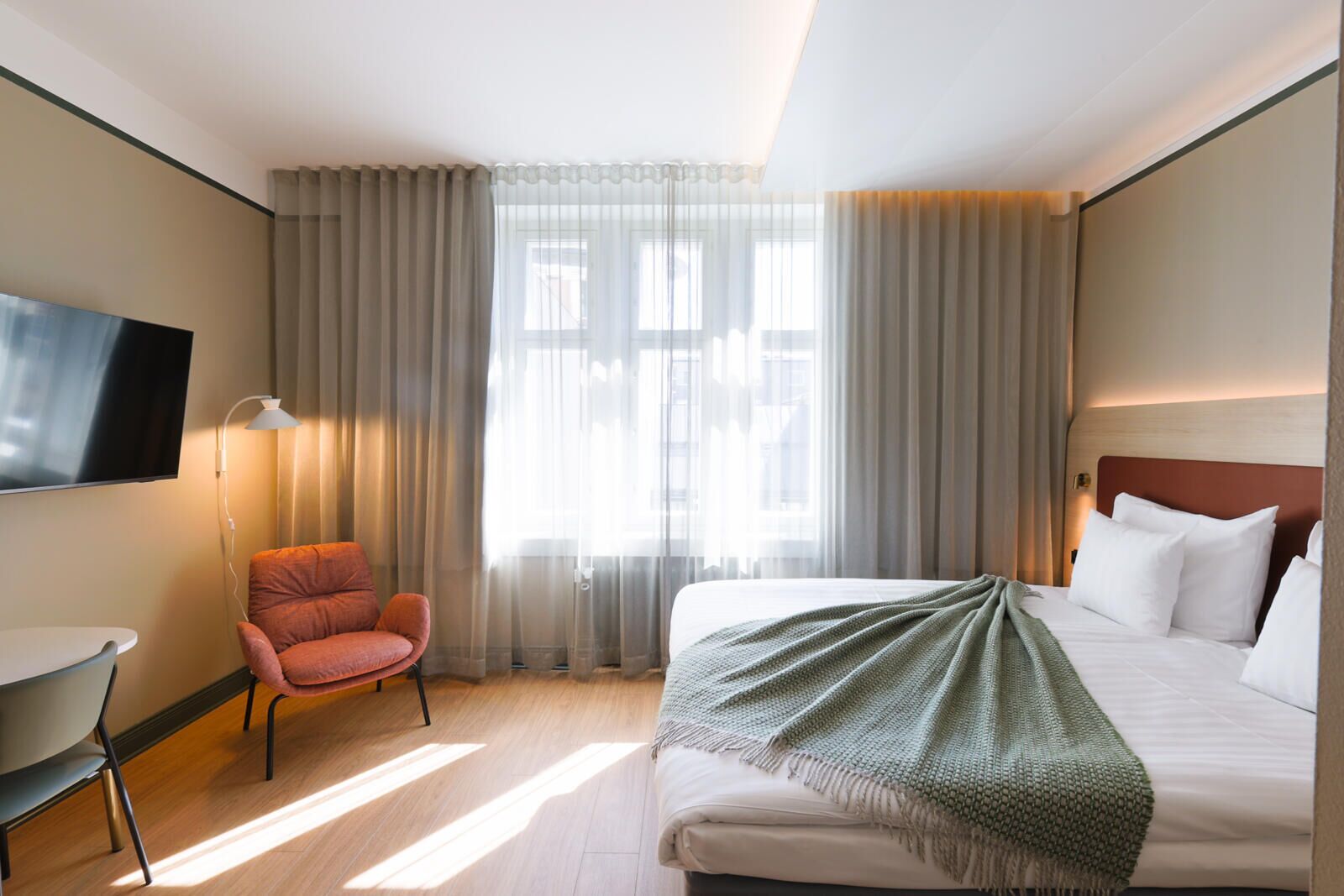 Une chambre d’hôtel avec un lit, une chaise, une télévision et une fenêtre avec des rideaux.