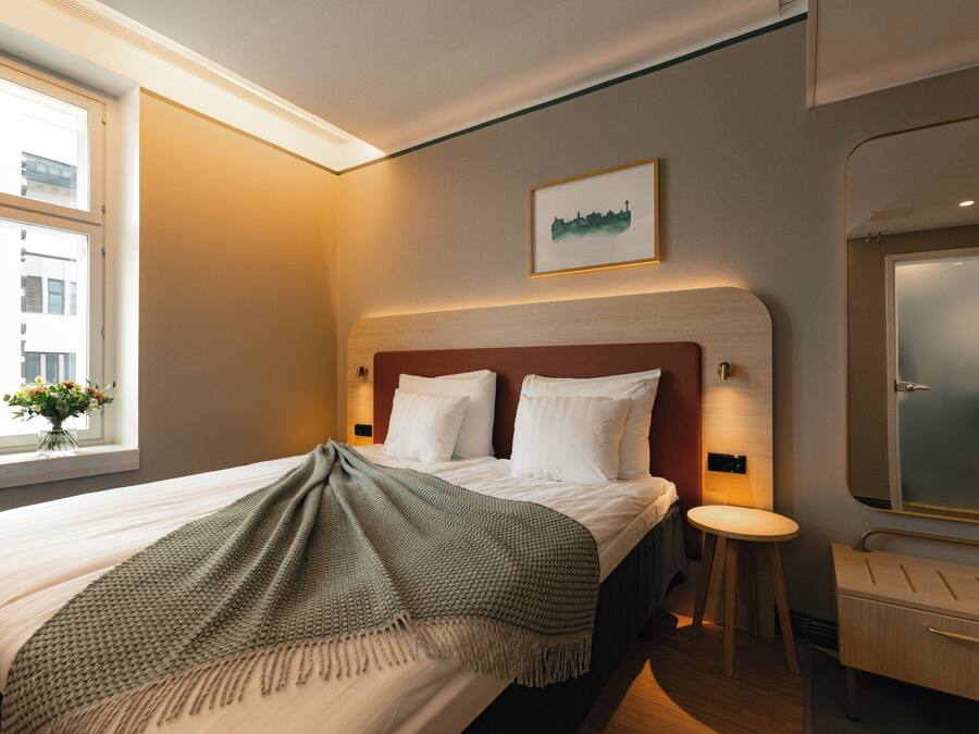 A hotel room with a bed, a bedside table, a mirror, and a window with a view of buildings.
