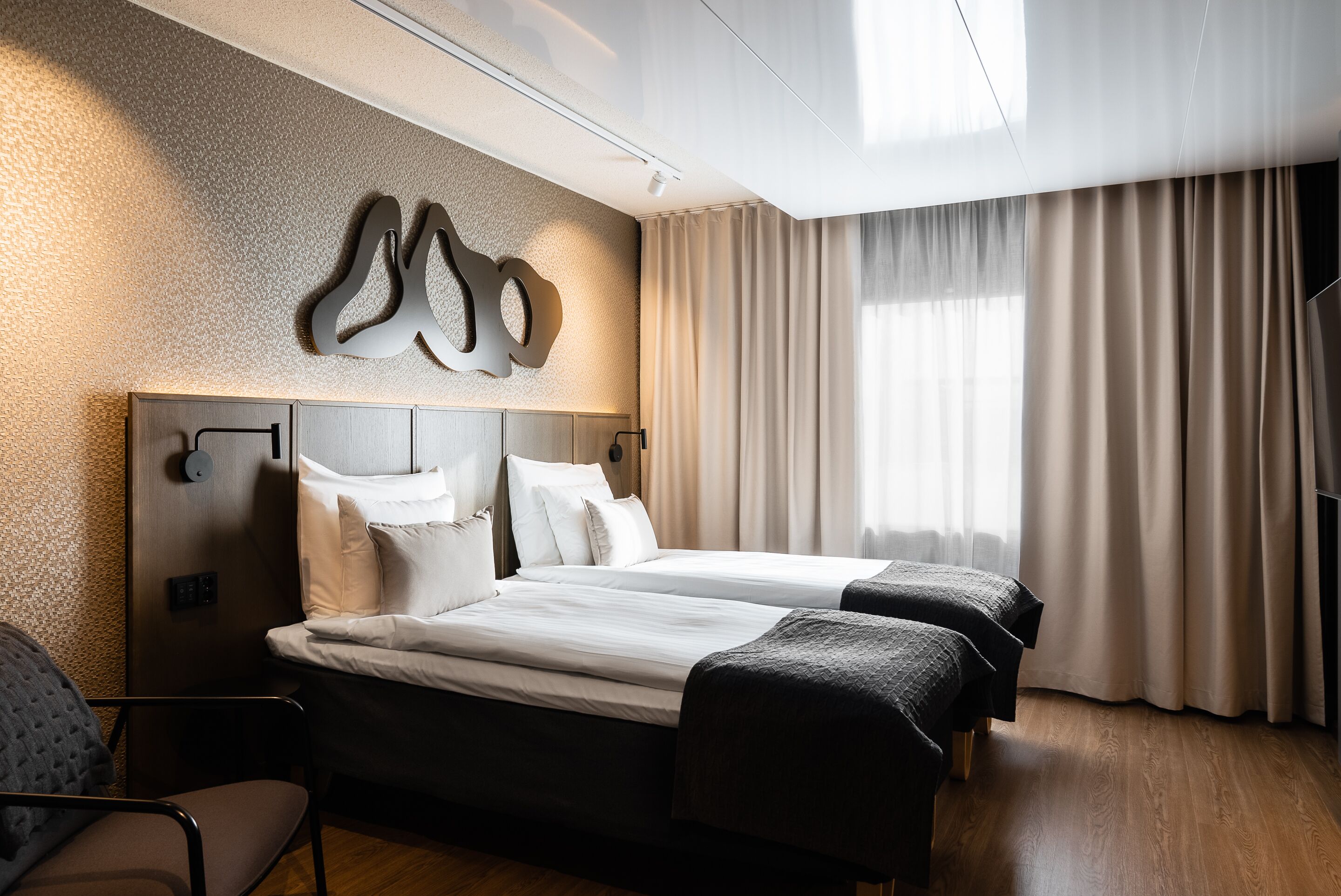 A hotel room with a large bed, two bedside tables, a headboard, a wall-mounted clock, and a large window with curtains.