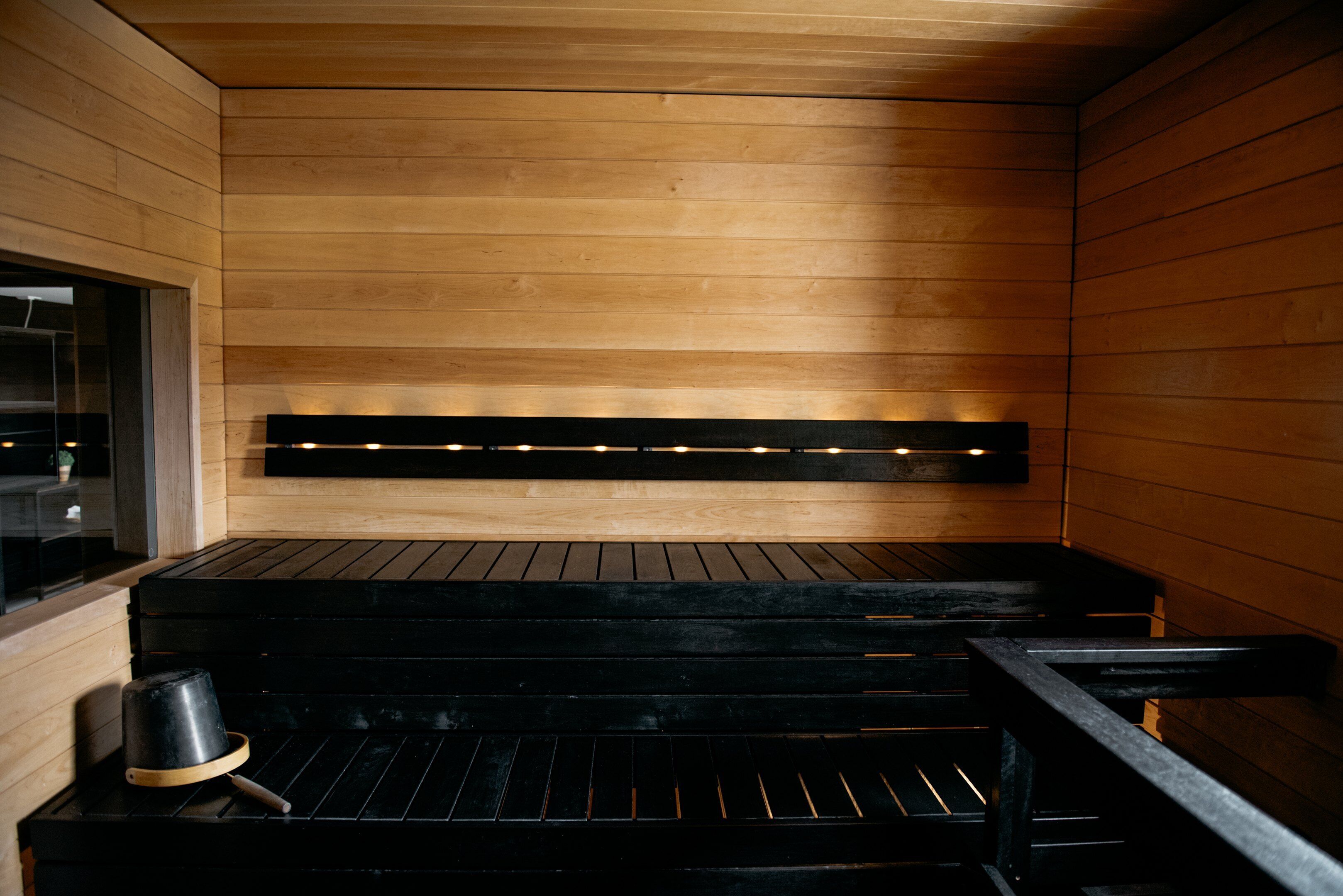 A sauna with wooden walls and benches, a bench with a bucket, and a lighted shelf above the benches.