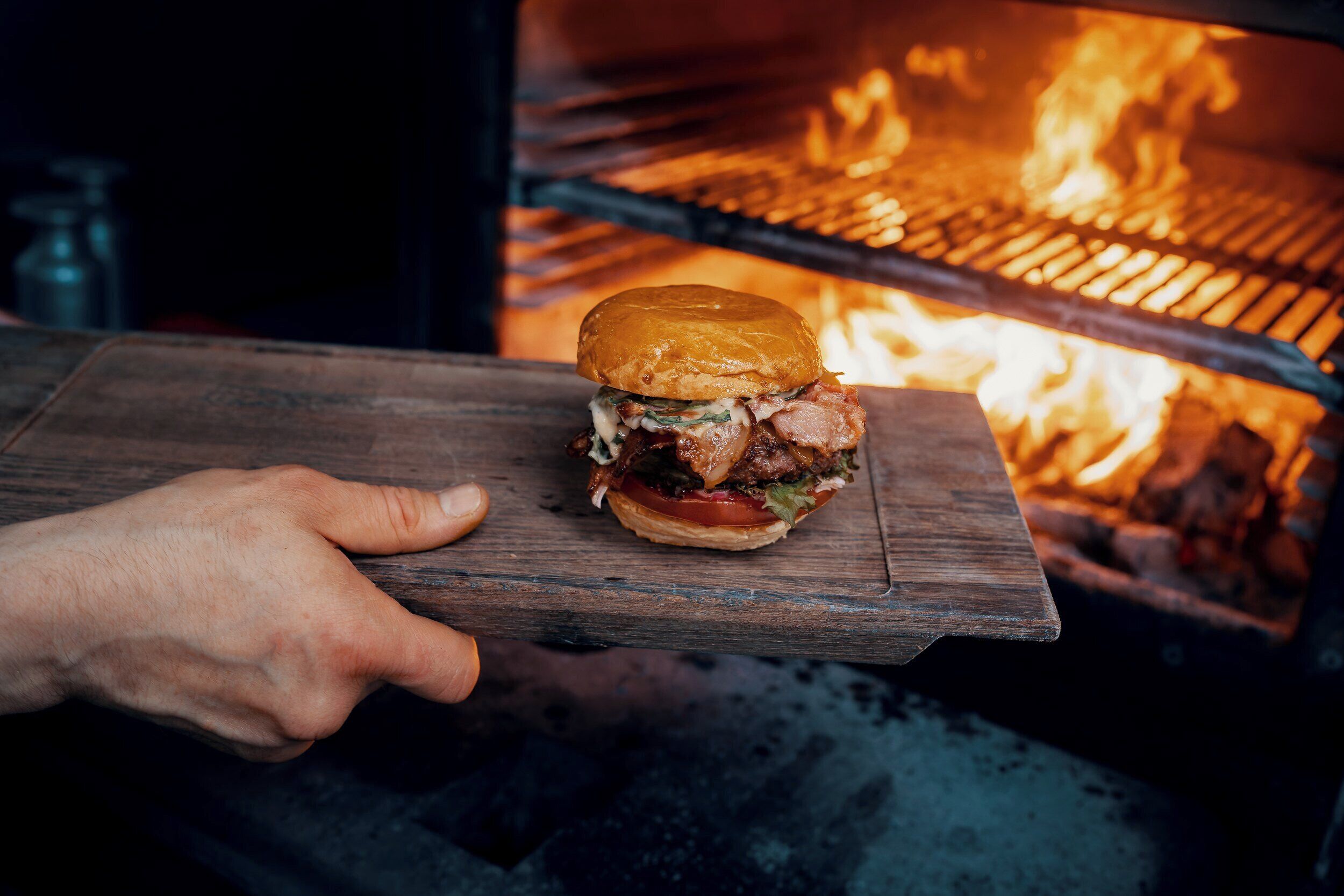 Una mano sostiene una tabla de madera con una hamburguesa frente a un horno de leña.