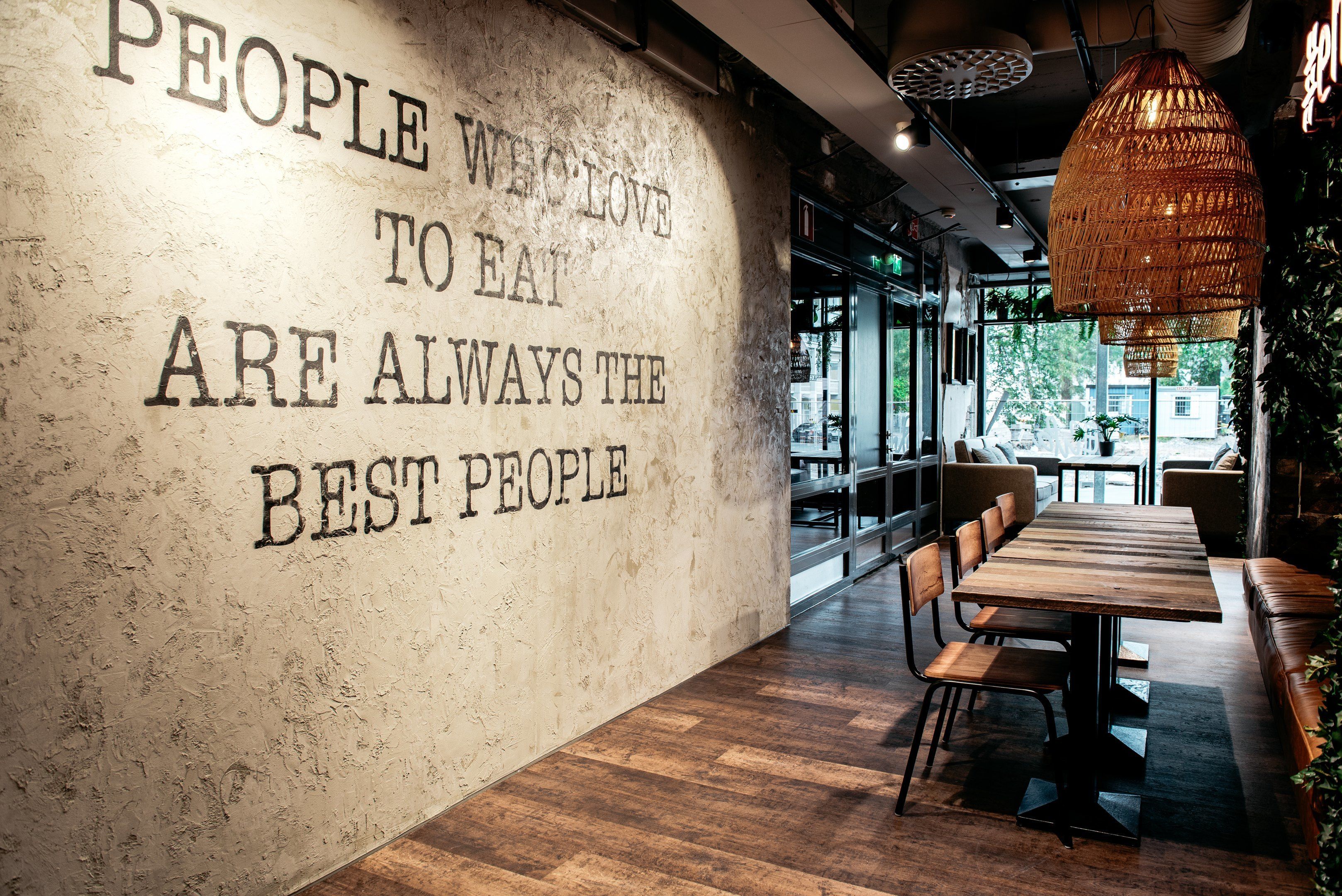 L’intérieur d’un restaurant, avec un grand texte mural, des tables et des chaises en bois, et un luminaire suspendu.