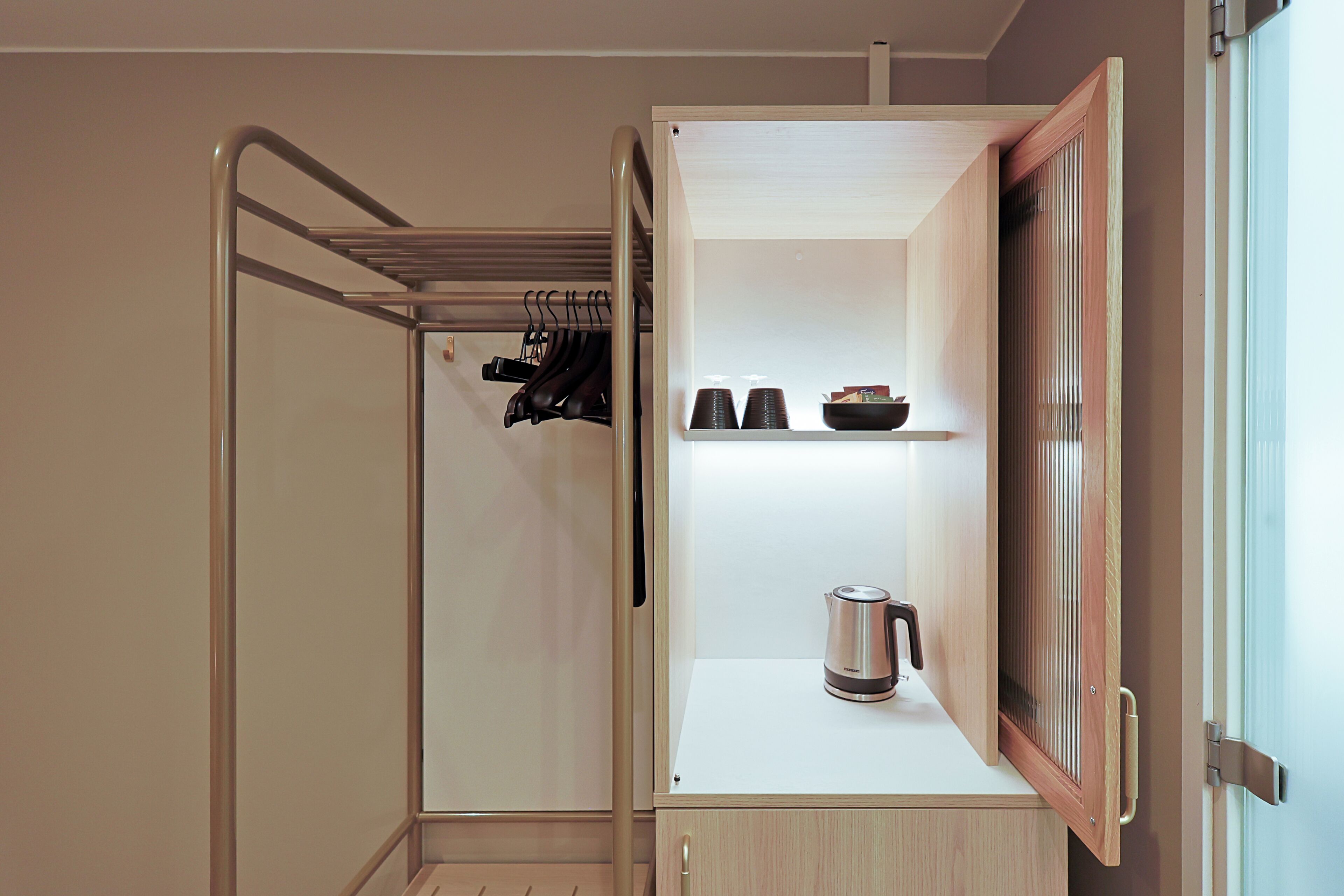 A compact room with a built-in wardrobe, a shelf with cups and a kettle, and a hanging rack with shoes.