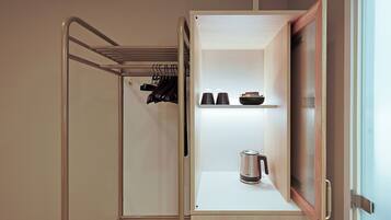 A compact room with a built-in wardrobe, a shelf with cups and a kettle, and a hanging rack with shoes.