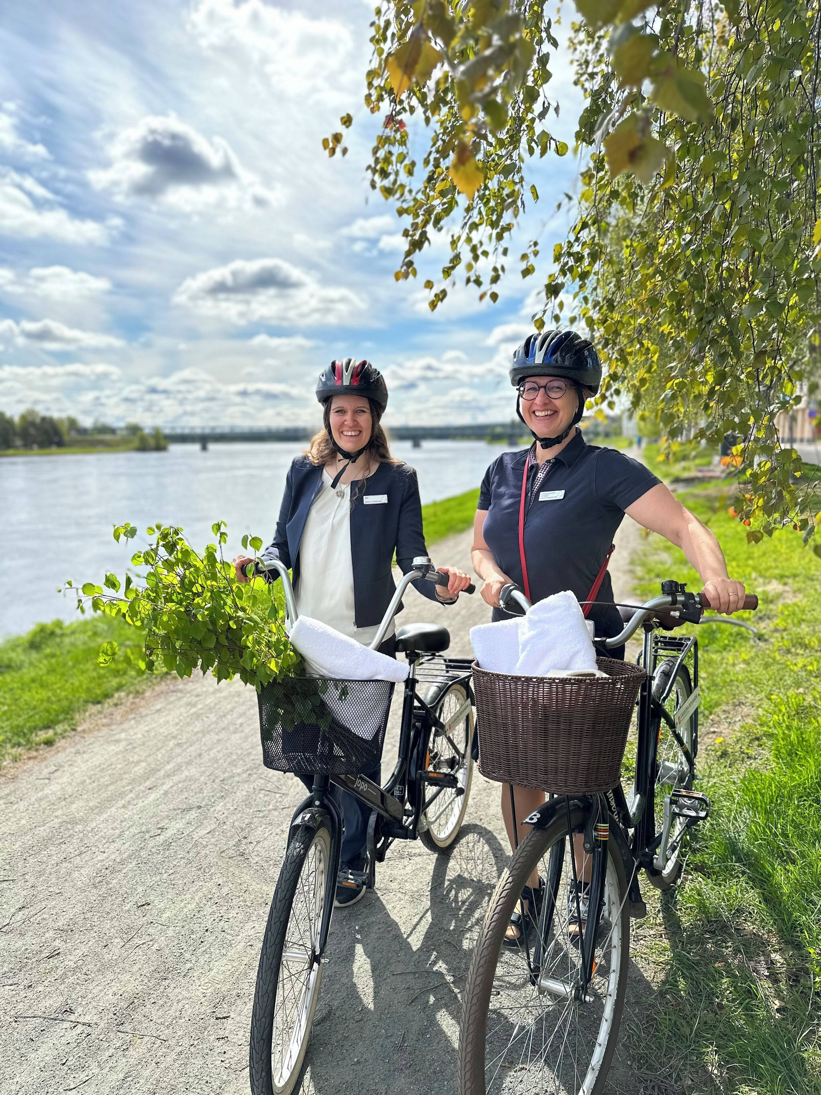 Twee mensen op fietsen met mandjes, een rivier op de achtergrond en een heldere lucht.
