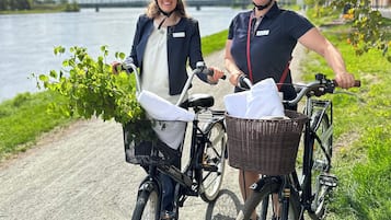 Twee mensen op fietsen met mandjes, een rivier op de achtergrond en een heldere lucht.