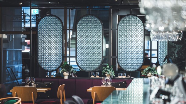 Un restaurante con una decoración moderna, que incluye un separador de vidrio con diseño de rombos, mesas de madera y asientos acolchados.
