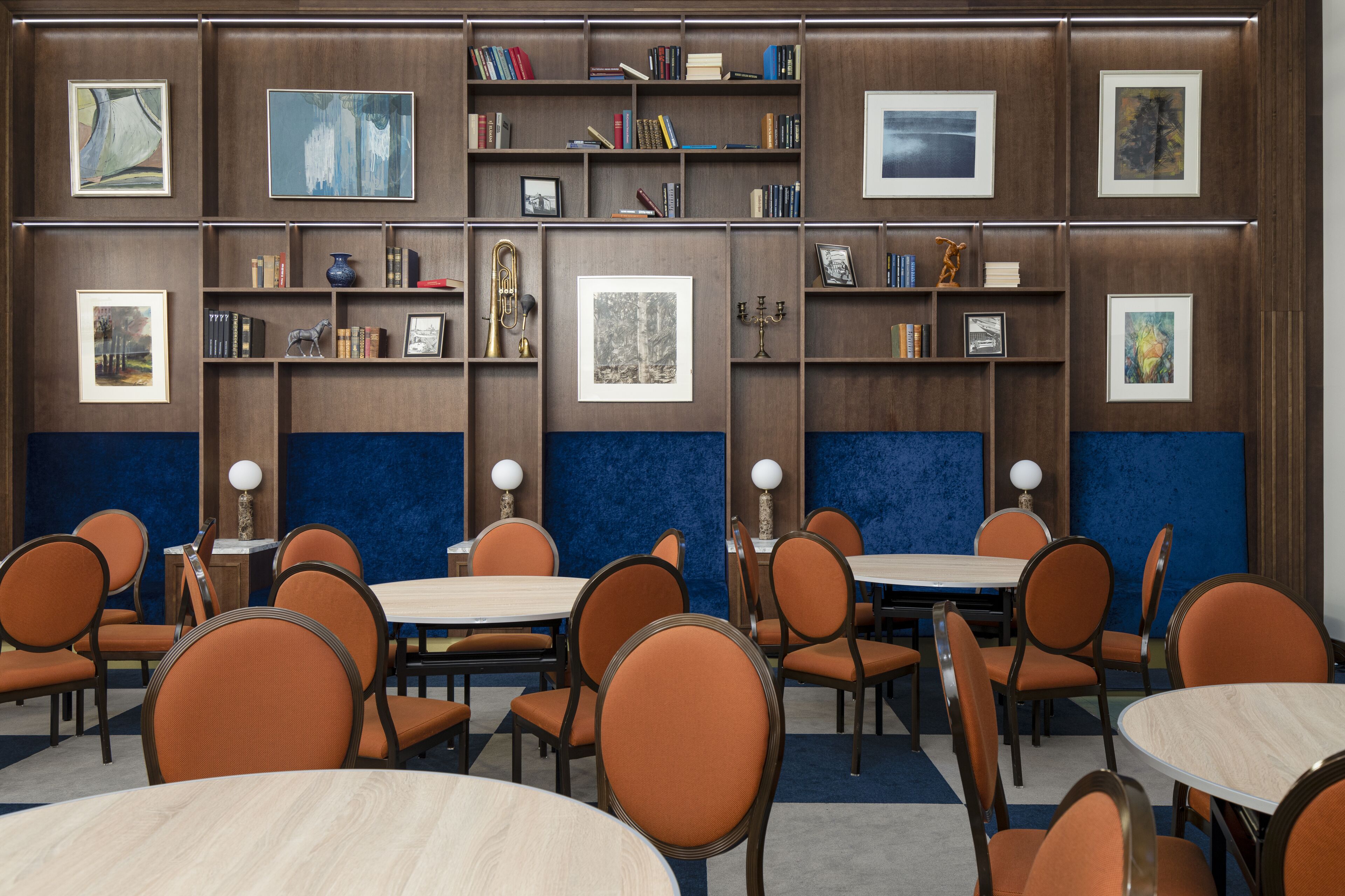 Un comedor con mesas redondas y sillas anaranjadas, rodeado de estanterías y cuadros enmarcados.