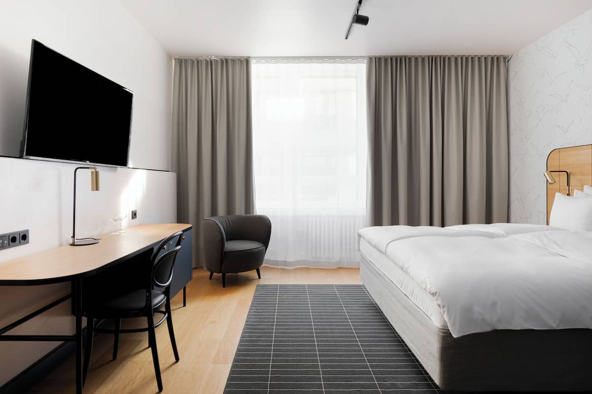 Una habitación de hotel moderna con una cama, un escritorio, una silla, un televisor de pantalla plana y un ventanal con cortinas.