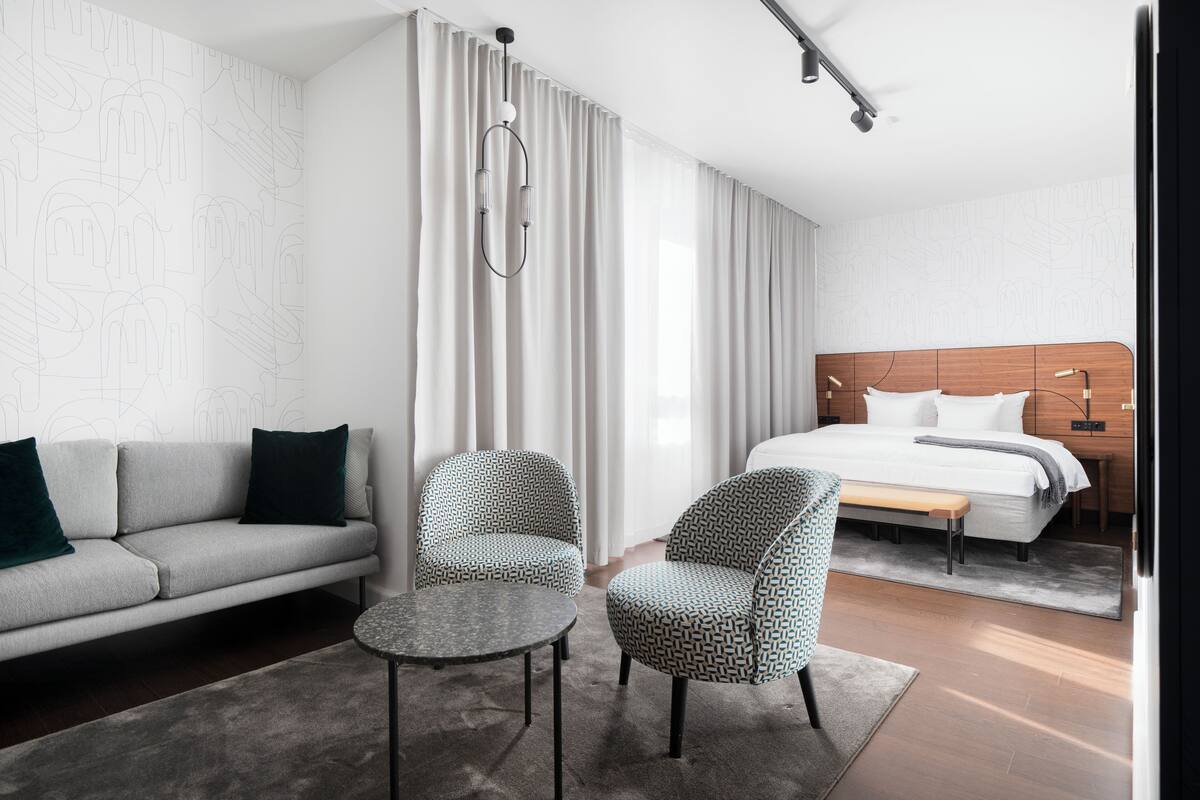 Una habitación de hotel moderna con sofá, dos sillas estampadas, una mesita, una cama con cabecero de madera y un ventanal con cortinas.