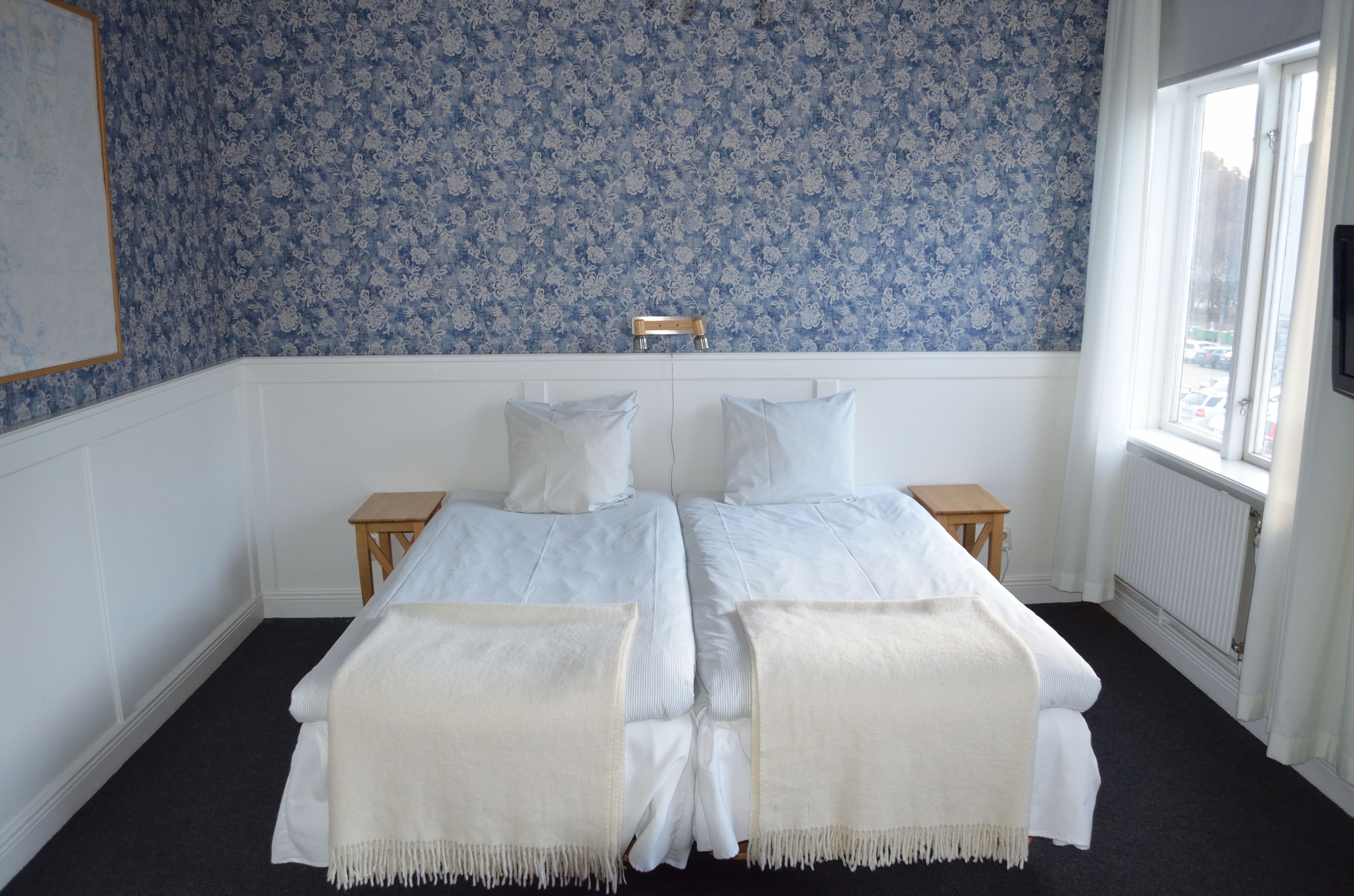 A double bed with white linens, two bedside tables, a wooden stool, and a window with a view.