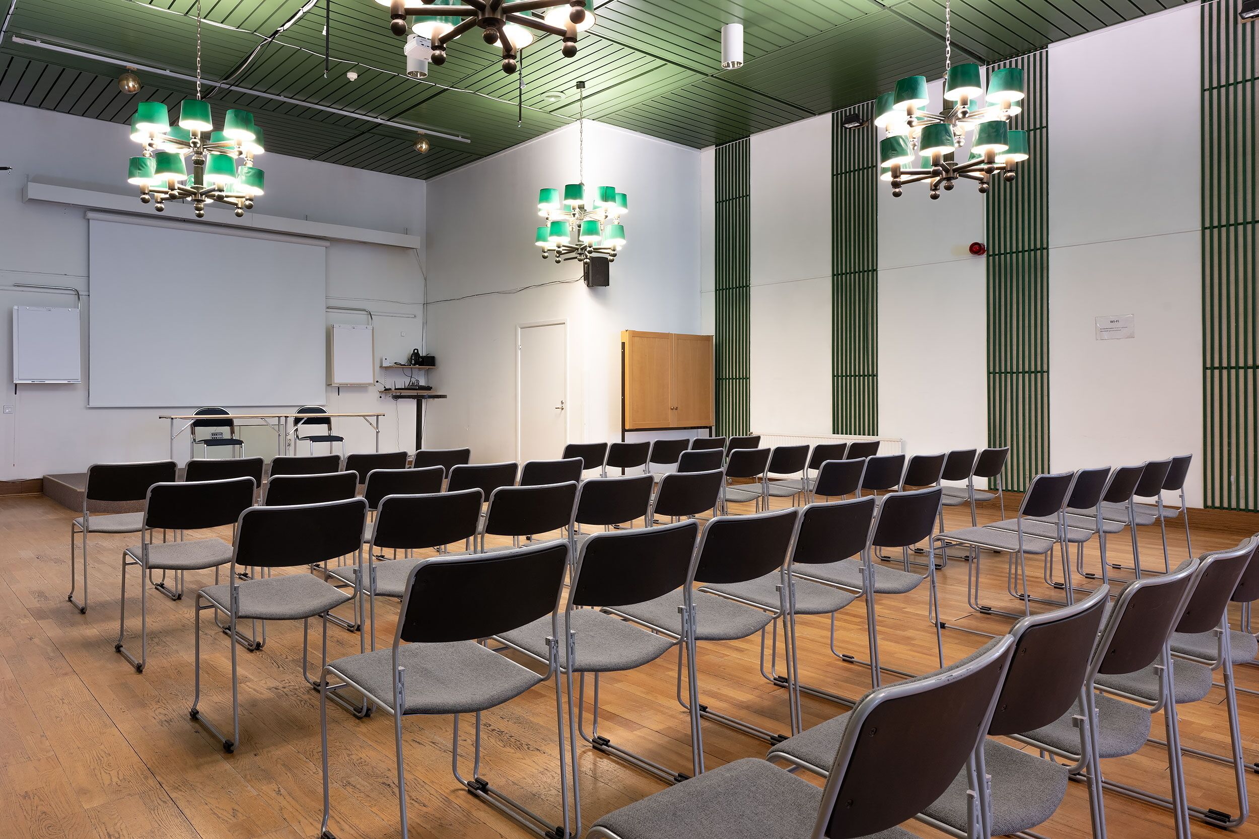 Una sala conferenze con file di sedie, uno schermo per proiettore e due lampadari.