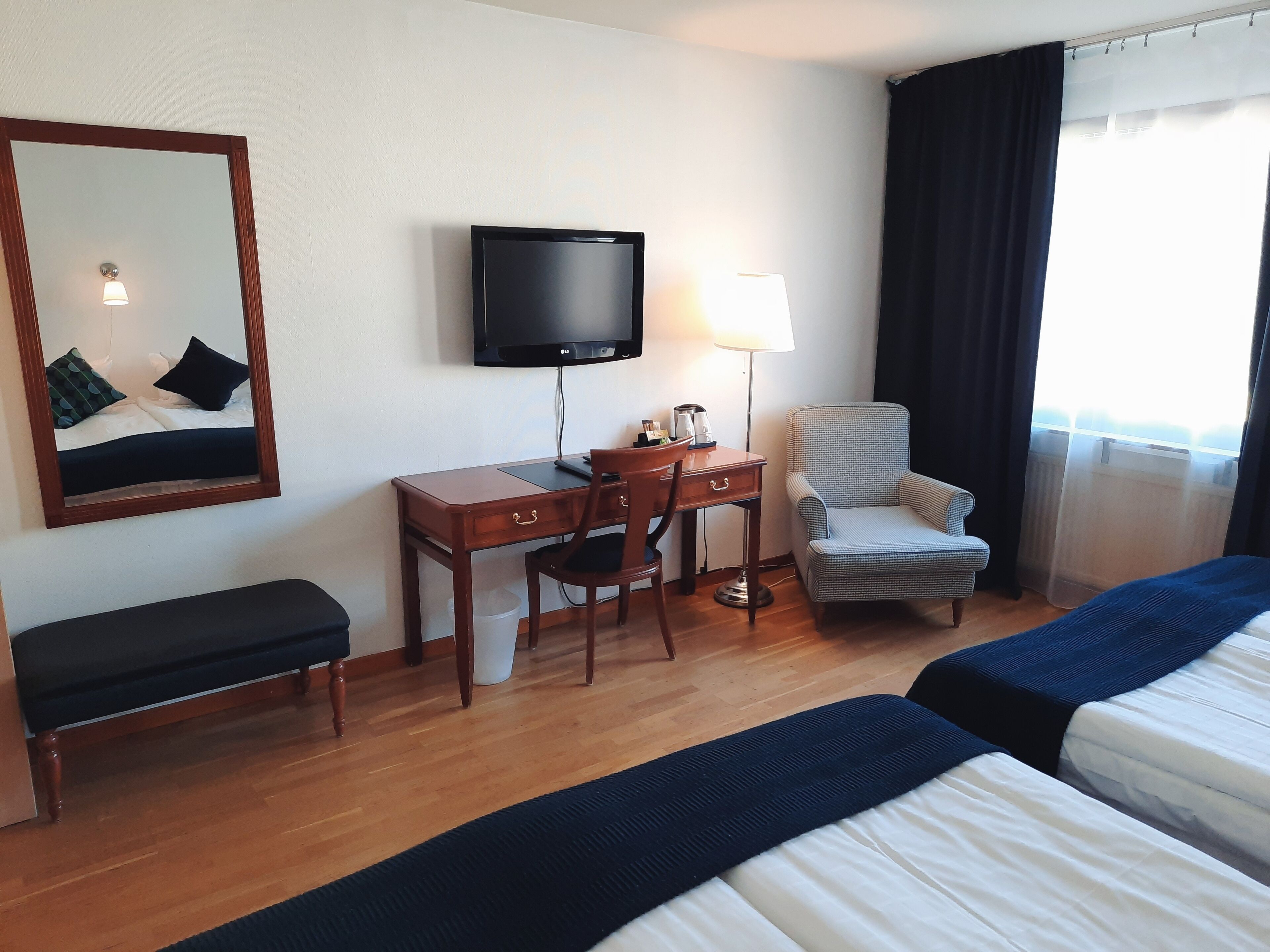 Habitación de hotel con una cama, un escritorio con silla, televisión, lámpara y ventana con cortinas.