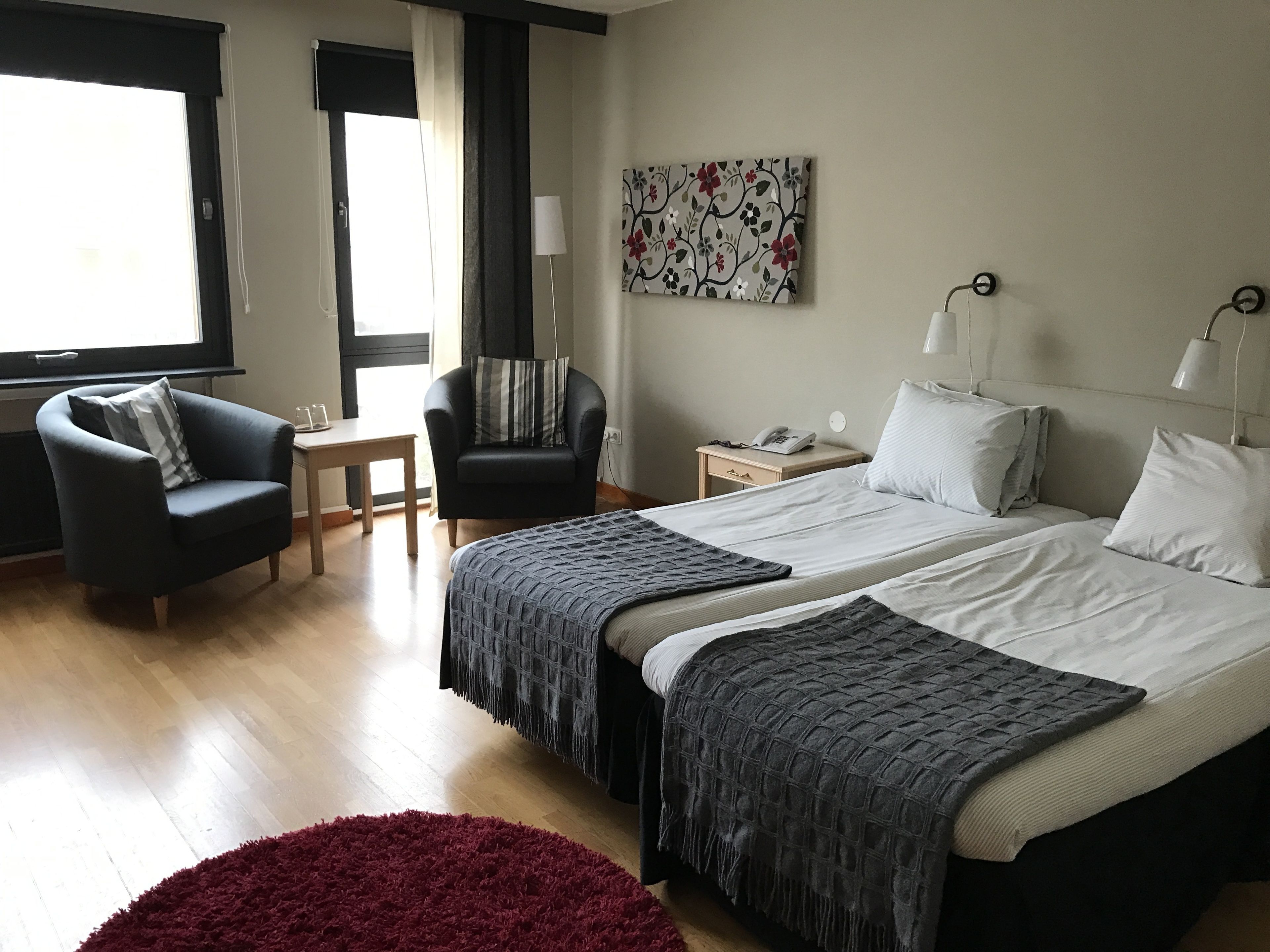 Habitación de hotel con dos camas, un escritorio, una silla, un tapete rojo y una ventana con cortinas.