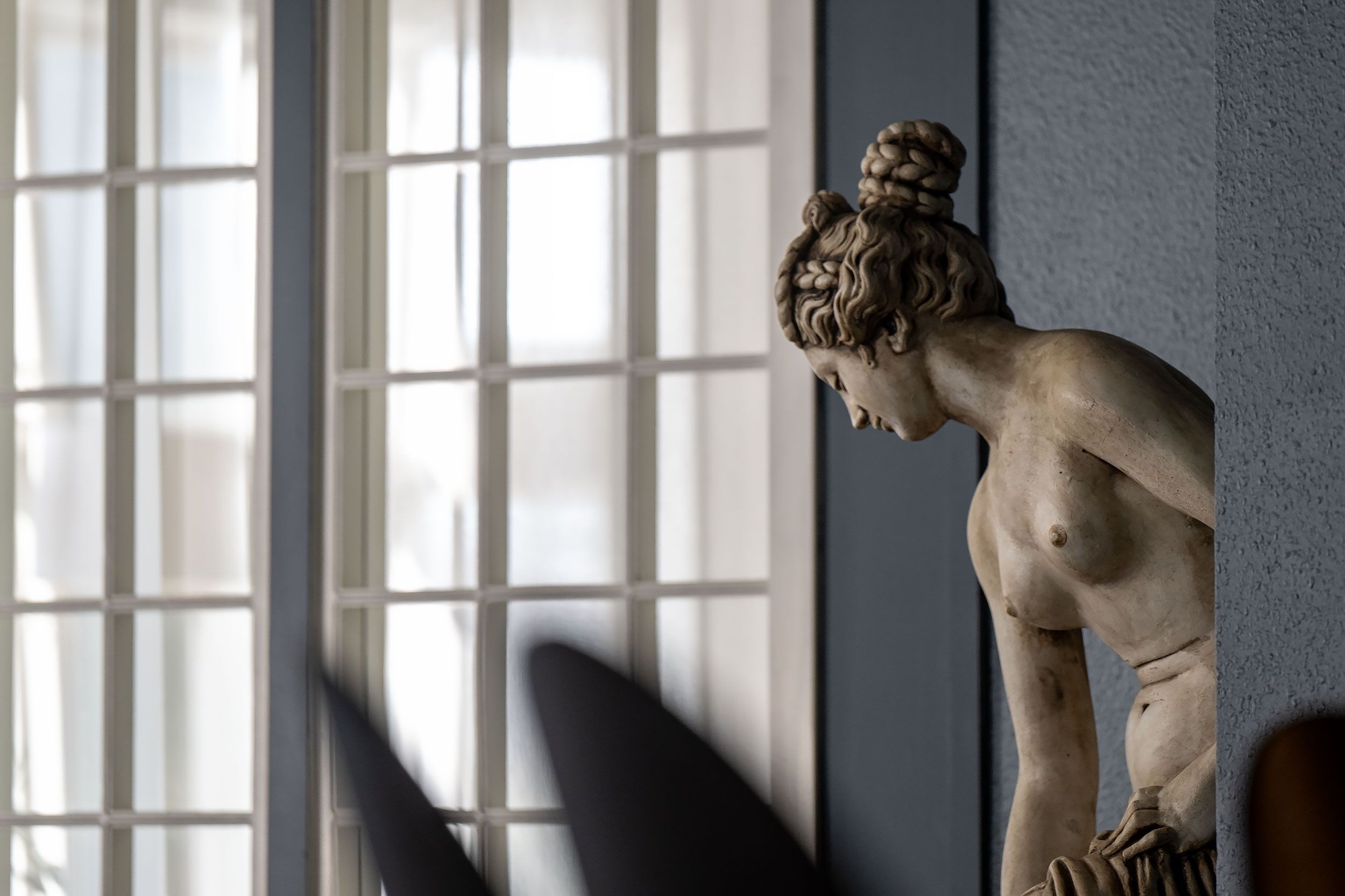 Une statue de marbre représentant une femme coiffée d’un chignon, placée devant un mur avec une fenêtre en arrière-plan.