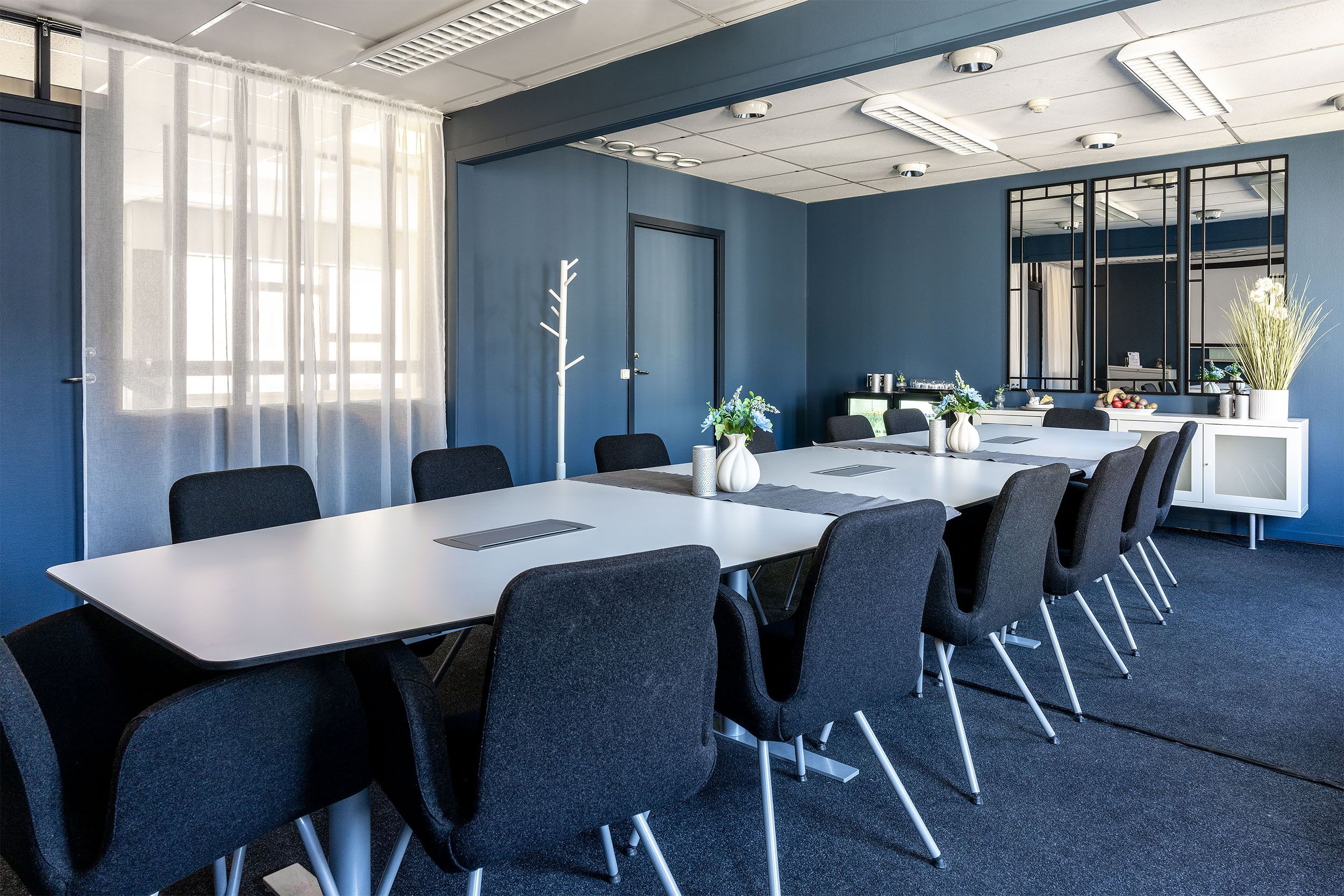 Une salle de conférence avec une longue table, des chaises noires, un tableau blanc et une plante en pot.