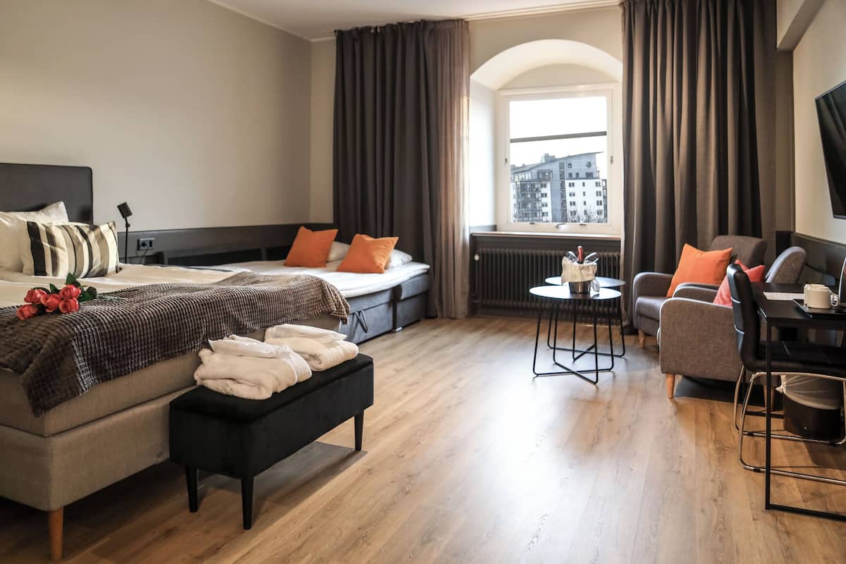 A modern hotel room with a large bed, a sofa, a small table, and a view of the city through a window.