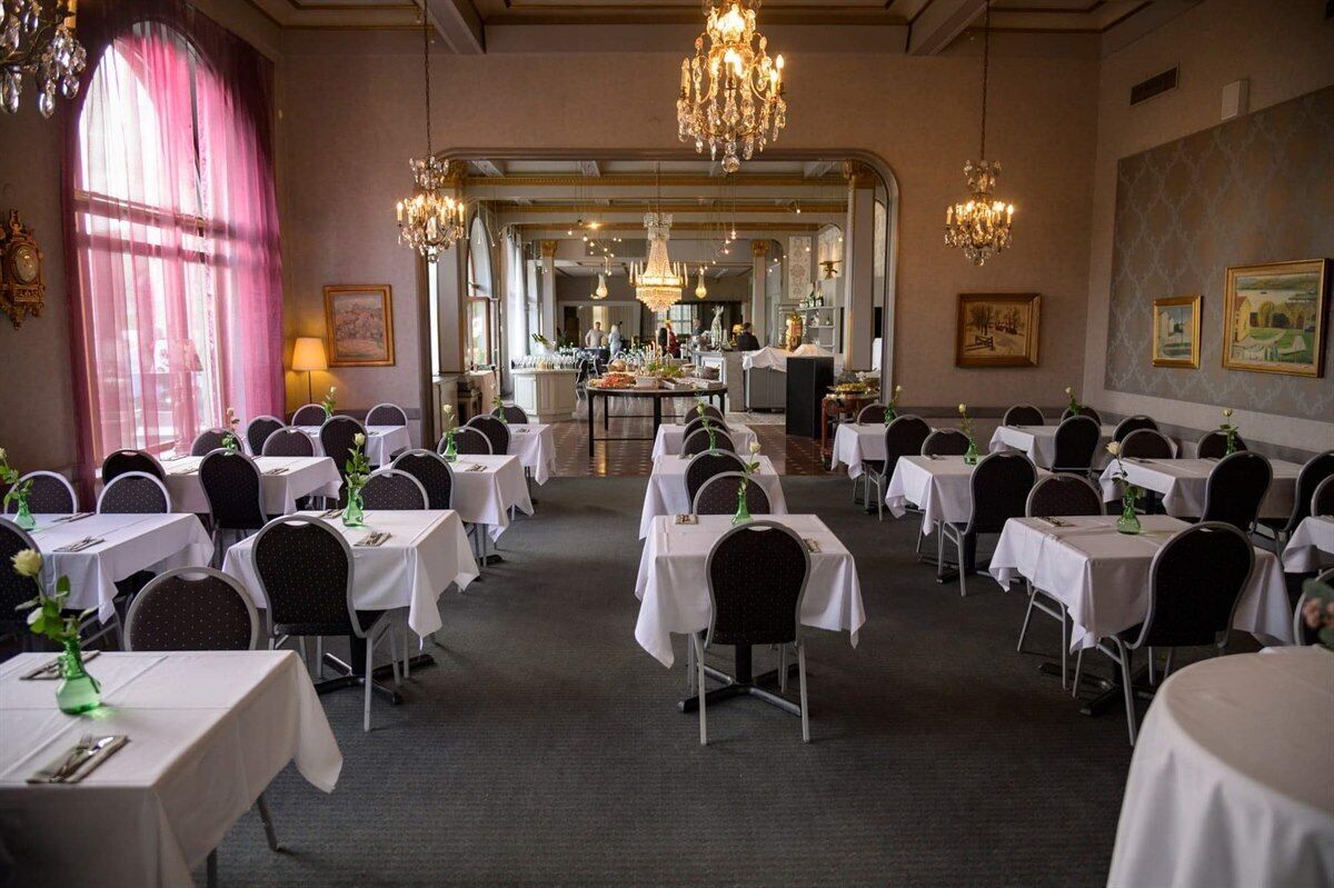 A formal dining room with multiple tables set for guests, chandeliers, and framed artwork on the walls.