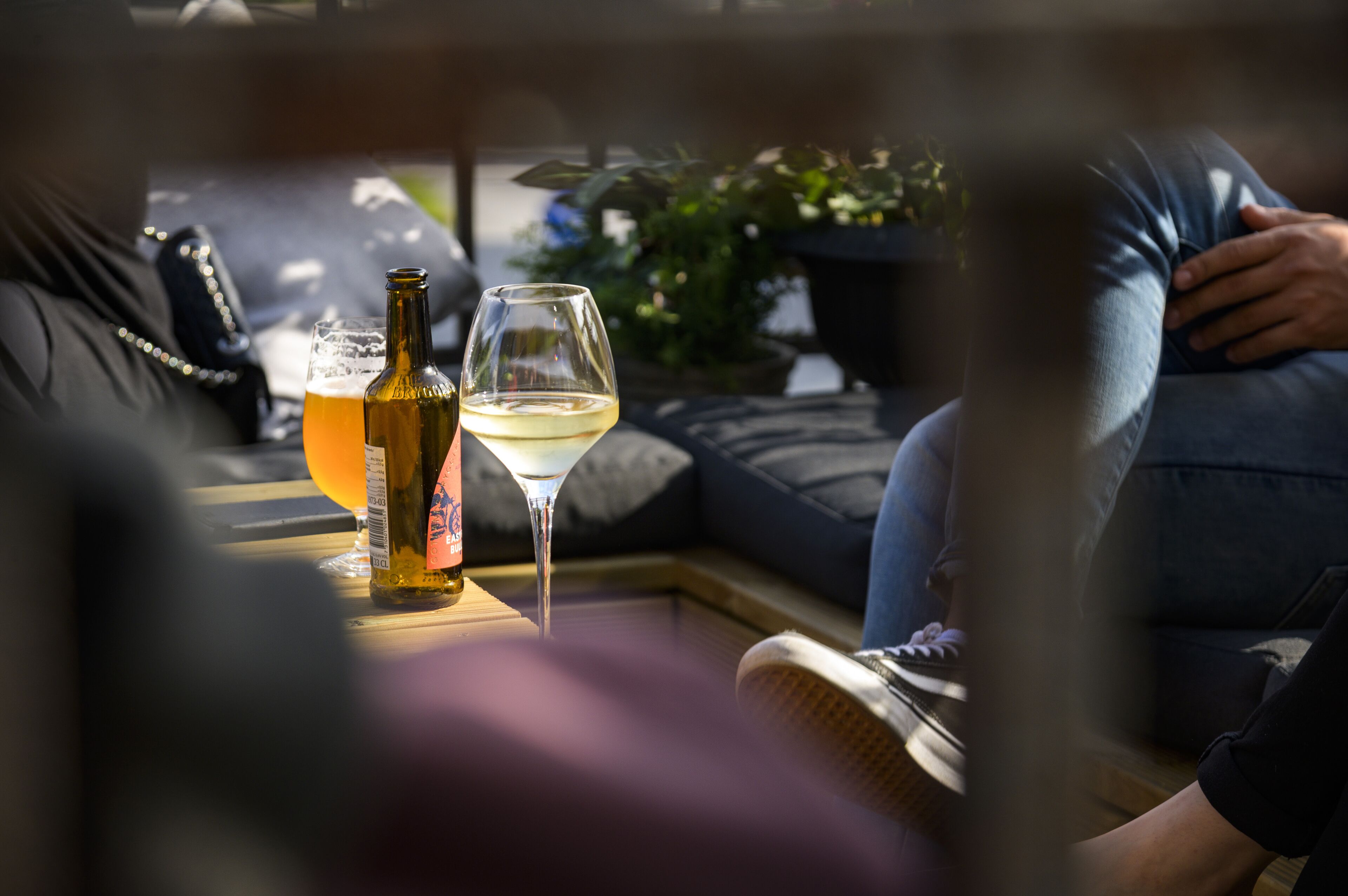 A table with a bottle and a glass of beer, a glass of white wine, and a person sitting on a couch.