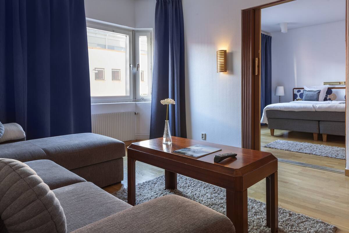 A modern hotel room with a sofa, a wooden table, and a view of the city through a window.