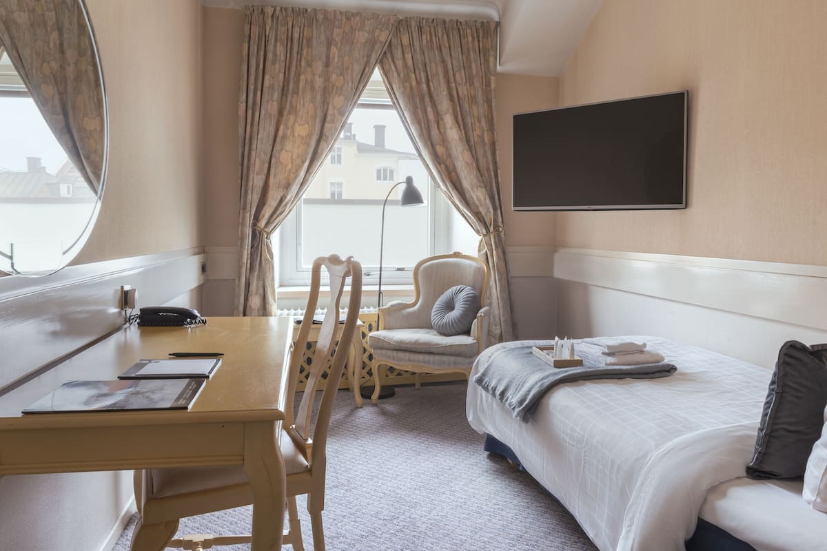 A hotel room with a bed, a desk with a chair, a flat-screen TV, and a window with curtains.