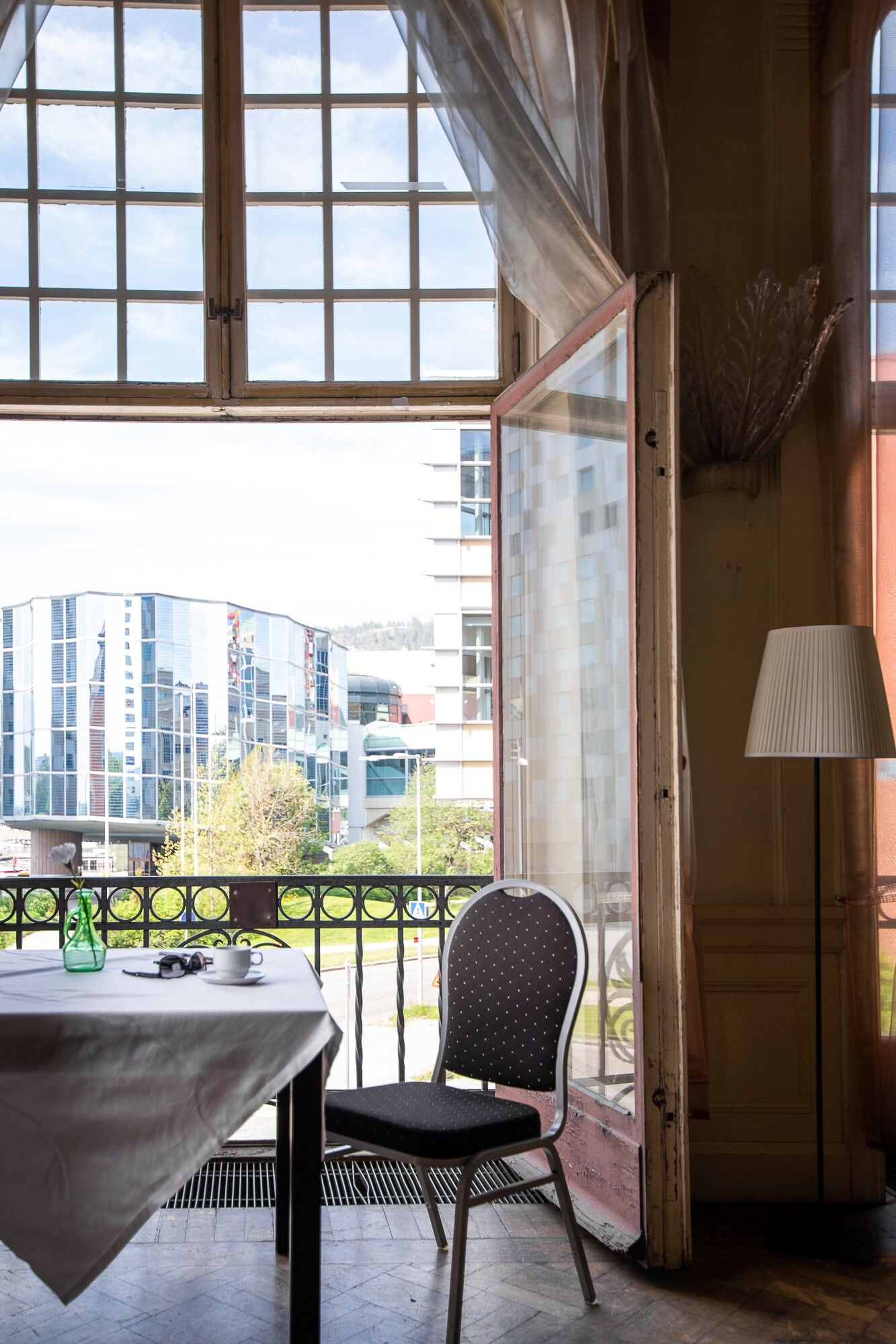 A room with a table and a chair, a lamp, and a view of modern buildings through large windows.