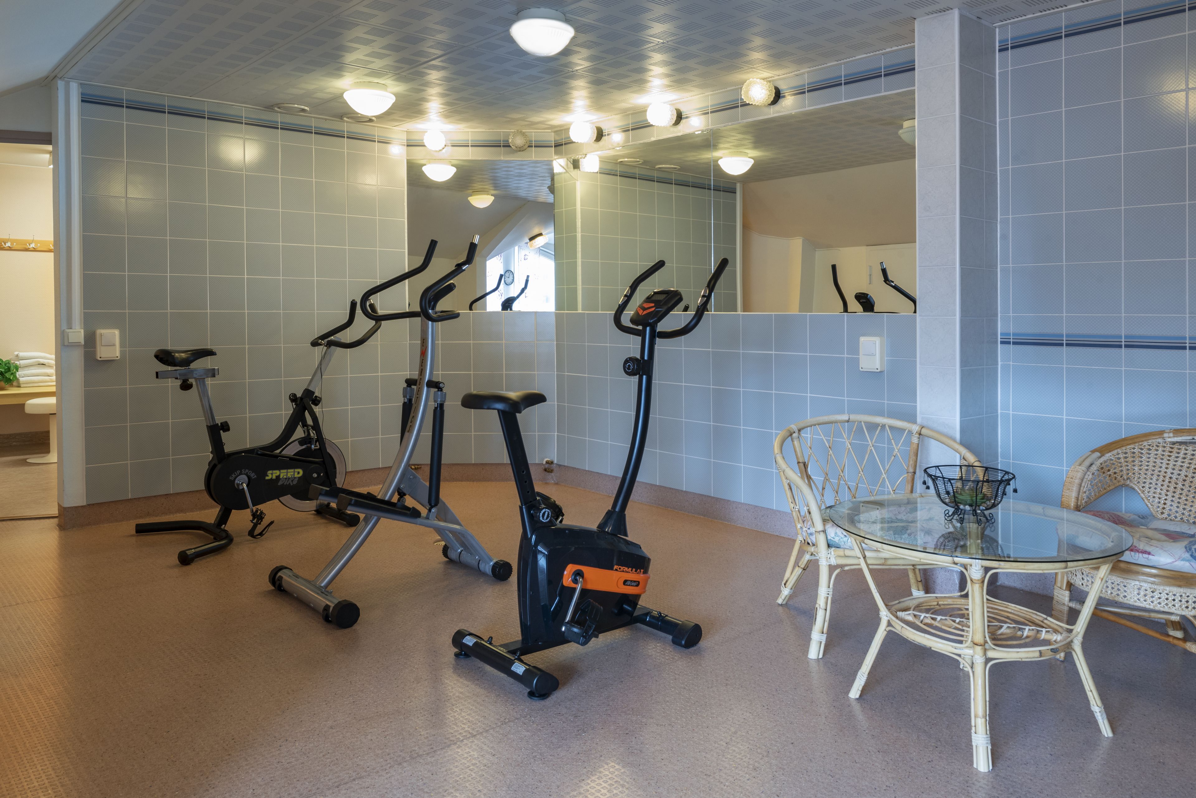 A room with three stationary bikes, a wicker chair, and a glass table.