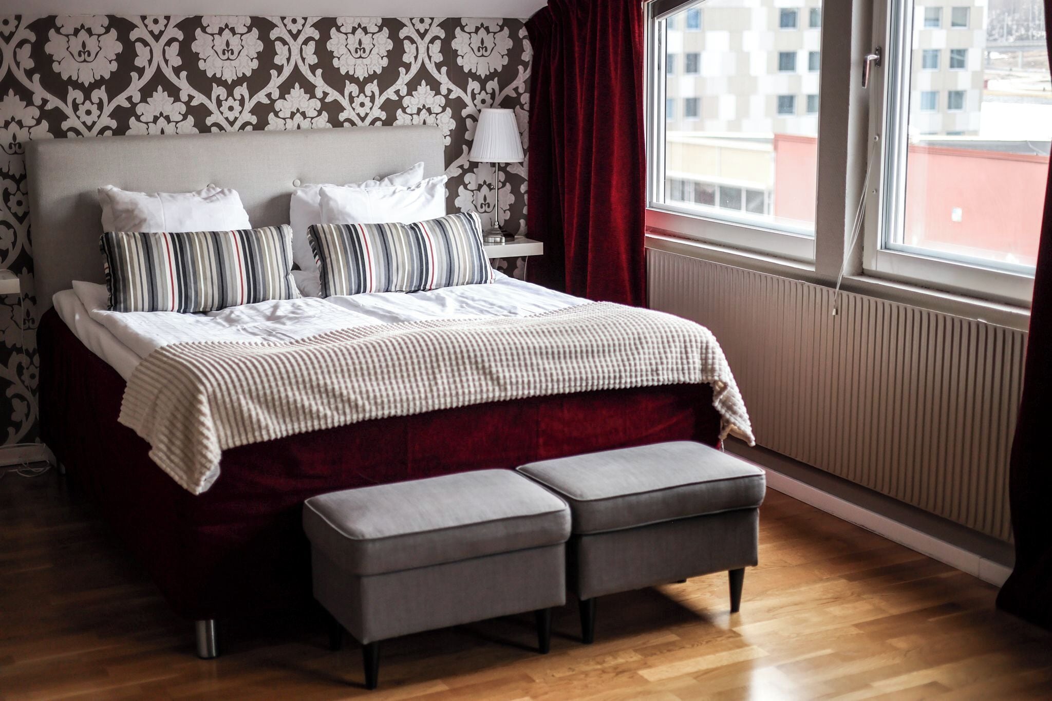 A hotel room with a large bed, a bedside table with a lamp, a window with curtains, and a view of buildings outside.