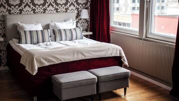 A hotel room with a large bed, a bedside table with a lamp, a window with curtains, and a view of buildings outside.