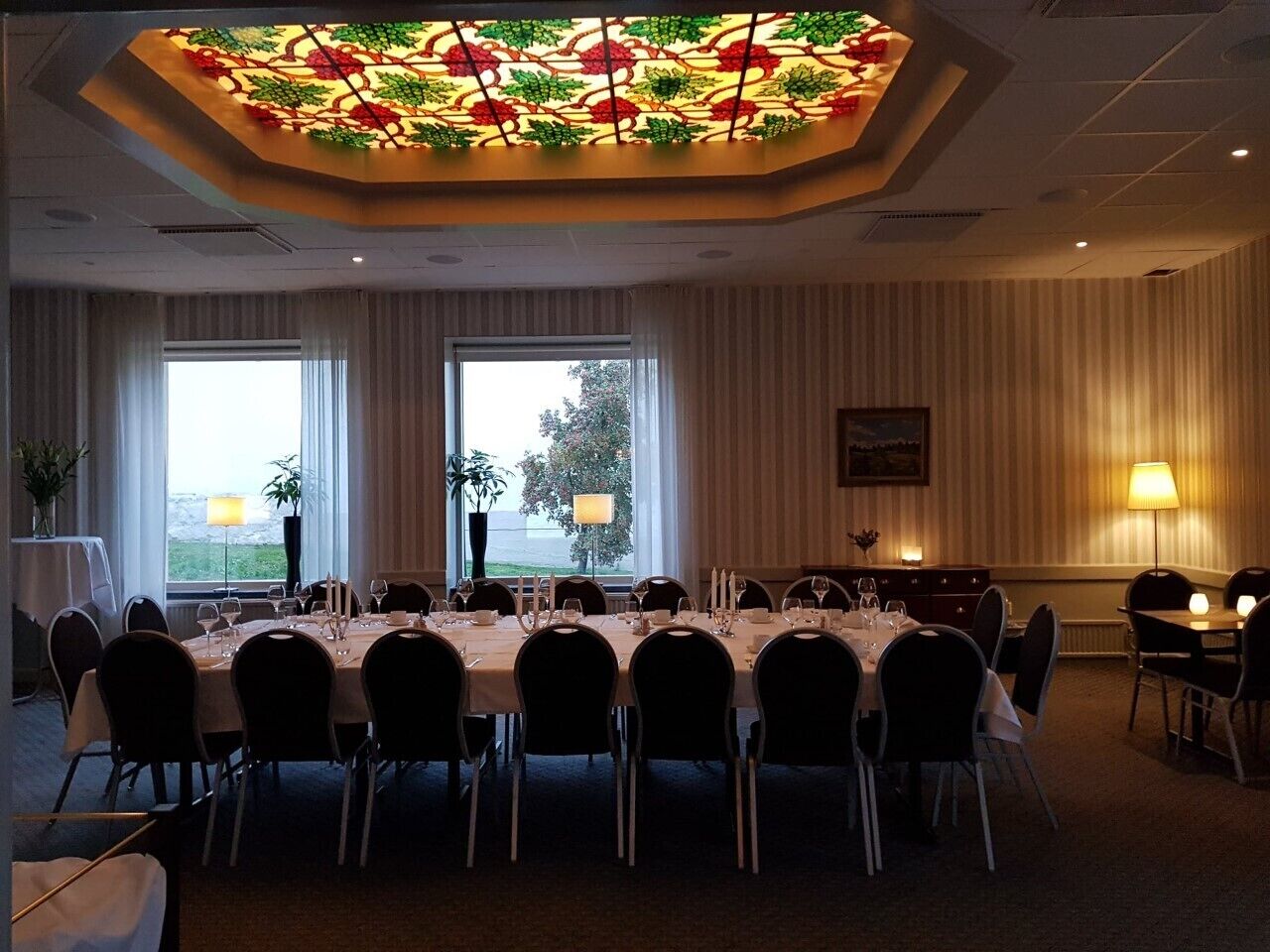 A conference room with a large table set for a meeting, two windows with curtains, a decorative ceiling light, and a lamp on the wall.